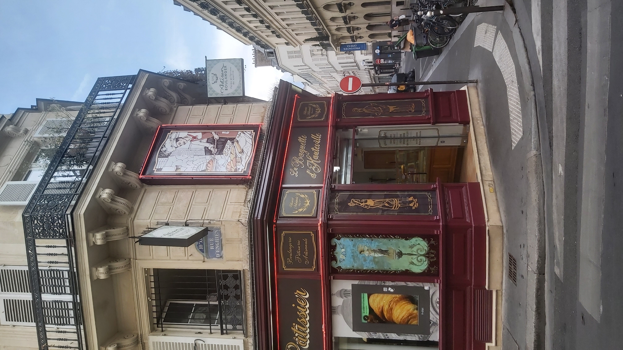 The image shows a corner of a building in Paris, France, with a bakery on the ground floor. The bakery has a dark red facade with gold lettering that reads "Pâtissier" on the left and "La Baguette d'Hauterville" on the right. There are also decorative panels with images of pastries and figures. Above the bakery, there is a sign that says "Artisan Boulanger." The building itself is made of stone and has traditional Parisian architectural elements, including shutters and a wrought-iron balcony. In the background, a street stretches out with more buildings and parked bicycles. A pedestrian crossing is visible in the foreground. The sky is overcast.