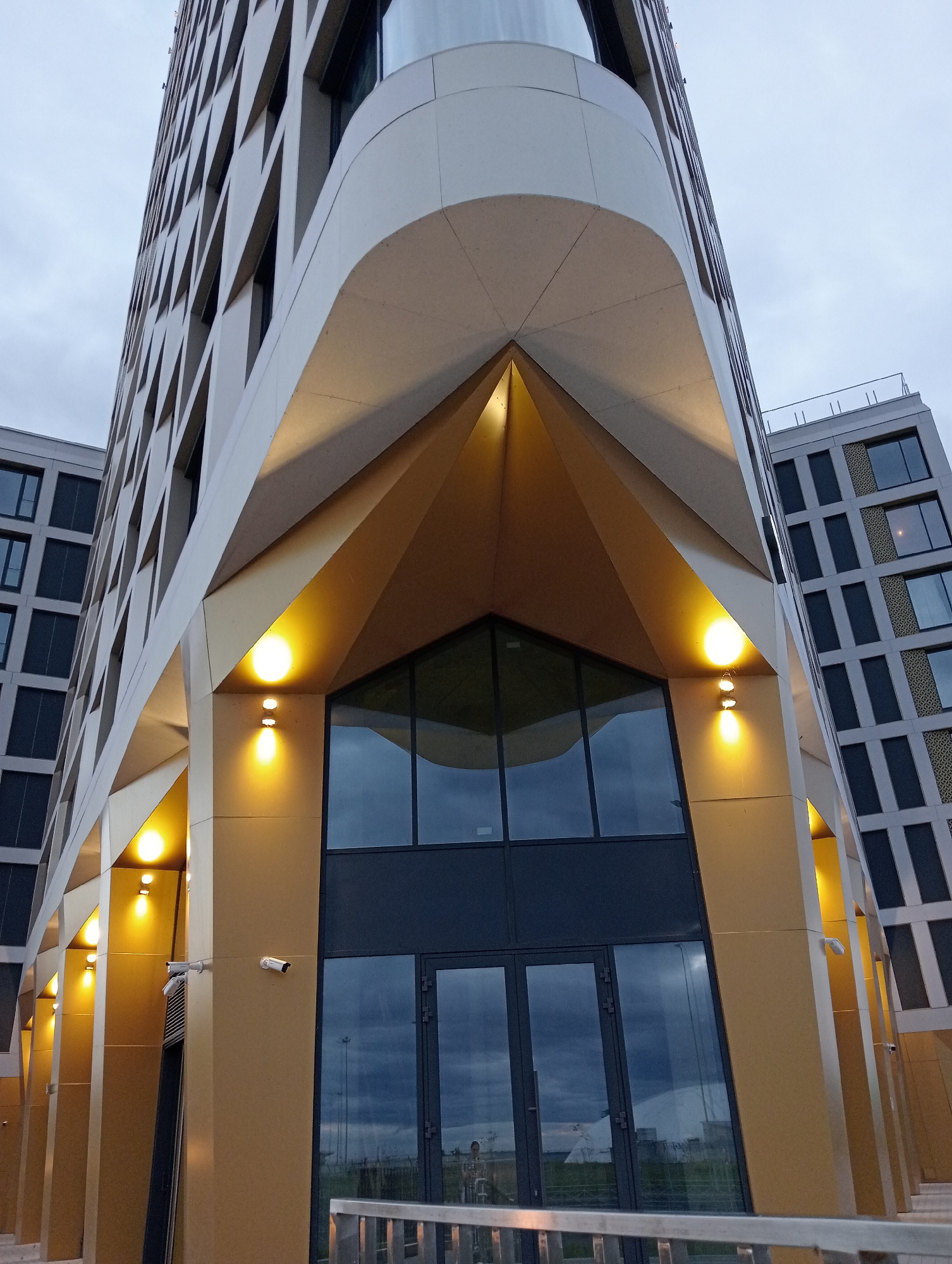 A low-angle, full-shot photograph captures the modern architectural design of a building located in Saint Petersburg, Russia. The building features a distinctive geometric facade with sharp angles and a predominantly light-colored exterior, accented by gold-colored cladding on the lower sections. Several yellow, circular exterior lights illuminate the entrance area, casting a warm glow. Two white security cameras are mounted on a pillar near the entrance. The building's entrance consists of large, dark-tinted glass doors. Adjacent to this structure, another building with a similar grid-like facade of dark windows and lighter accents is visible. The sky is overcast and a pale grey, suggesting a cloudy day. A railing made of metal bars is present in the foreground.