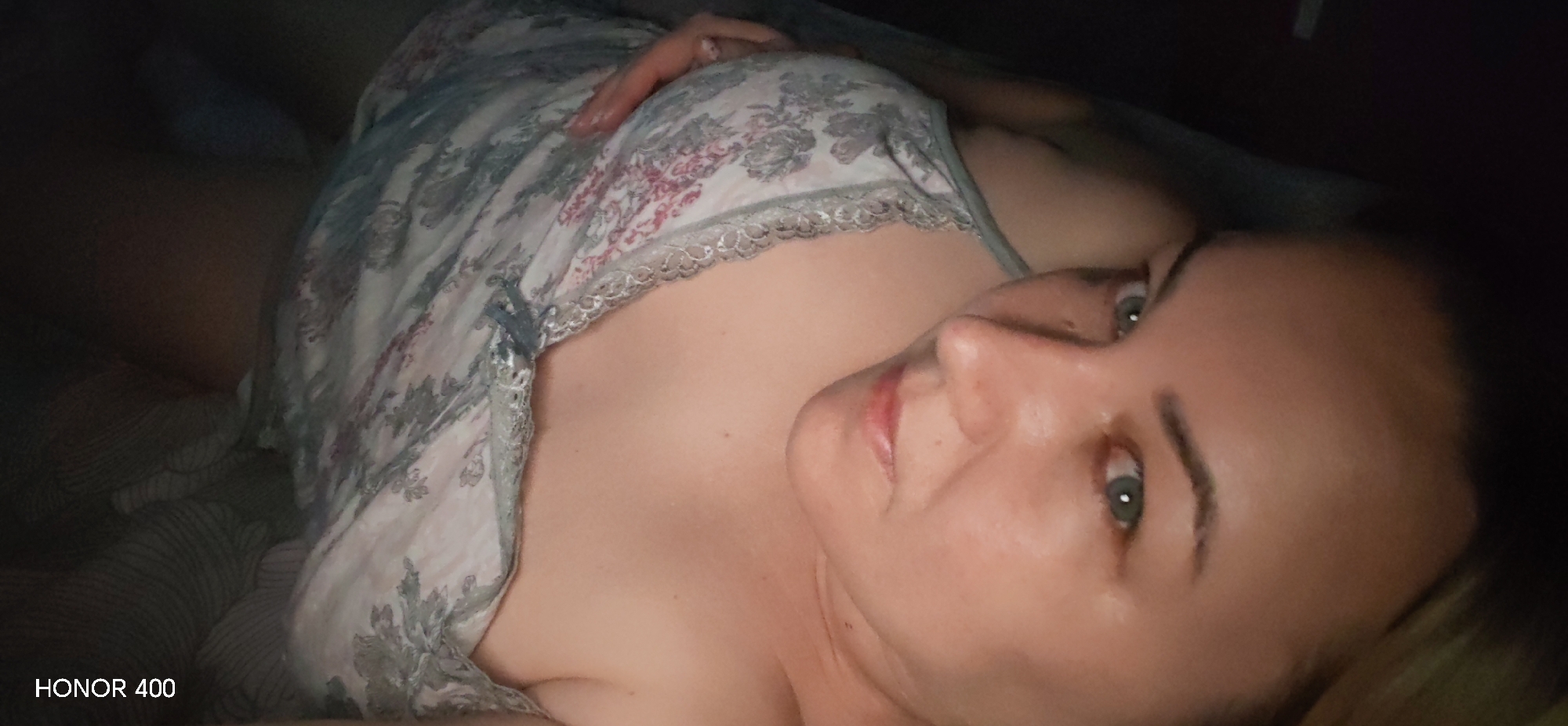 The image depicts a woman lying in bed. She is wearing a floral patterned nightgown with lace trim. Her head is turned towards the camera. The background is dark. The image includes the watermark "HONOR 400" in the bottom left corner. There is no indication this photo was taken in Noviy, Russia.