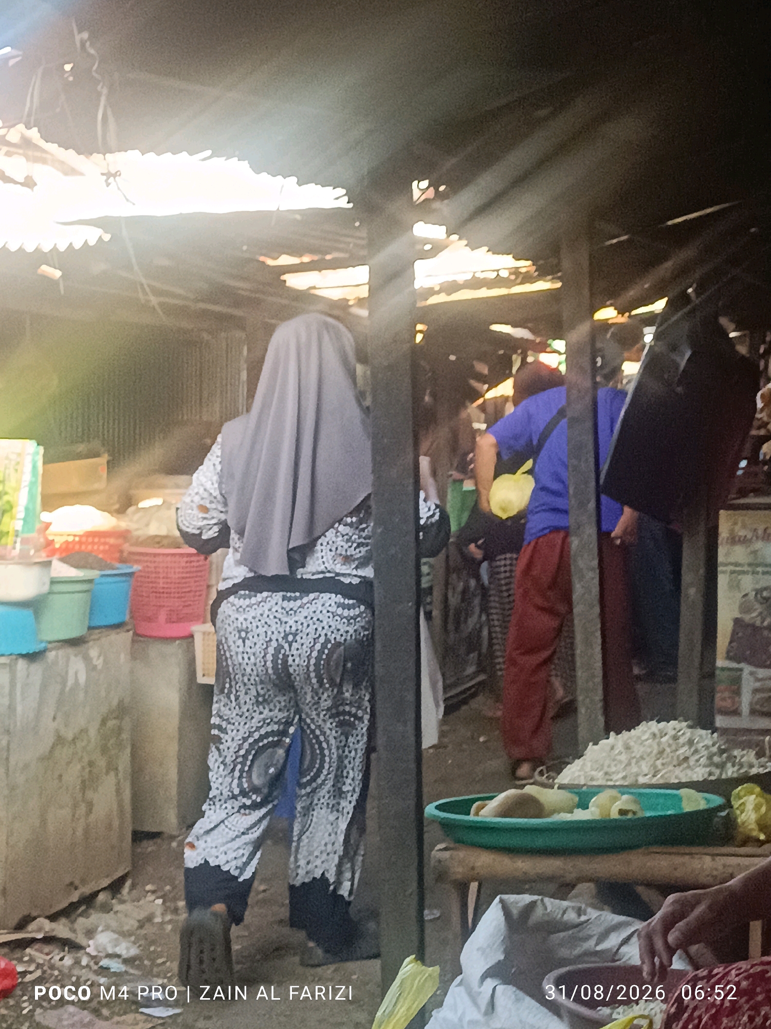 In the city of Arosbaya, Indonesia, a market scene unfolds under a partially covered structure. Sunlight streams through gaps in the corrugated metal roof, illuminating dust motes and casting long shadows. A woman, seen from the rear, wears a grey hijab and a black and white patterned outfit. She stands near a display of colorful plastic baskets containing unseen goods. To her right, another person in a blue shirt and red pants is partially visible. In the foreground, a green bowl holds light-colored food items. Bags and various market goods are scattered around the ground, suggesting ongoing commerce. A timestamp on the image reads "31/08/2026 06:52".