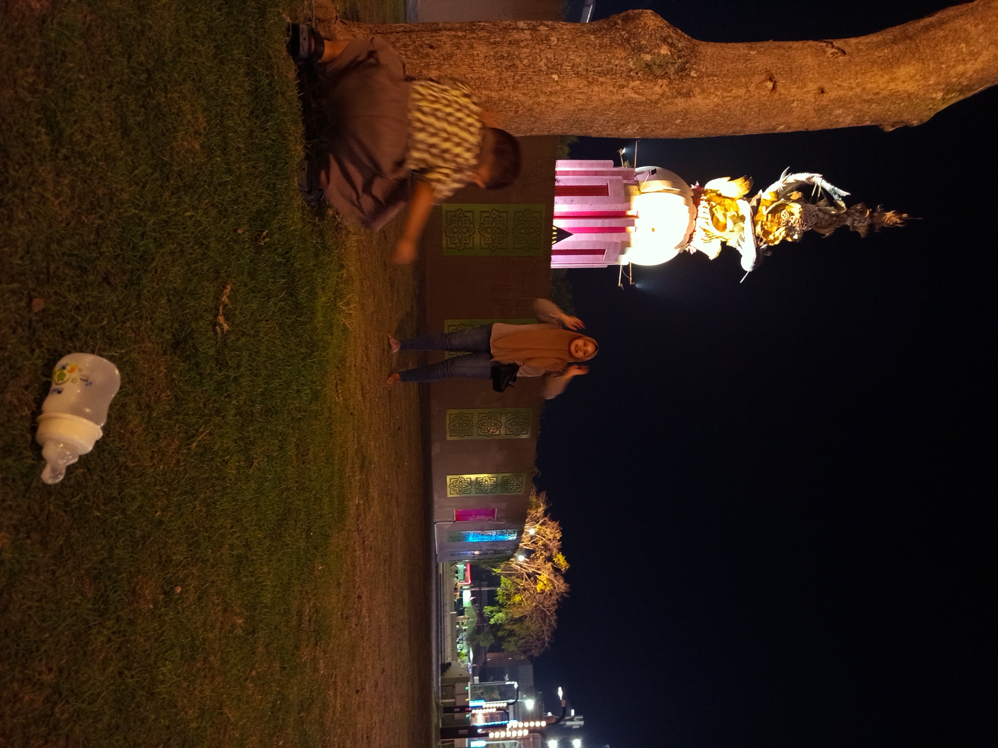 The image depicts a nighttime scene in a park or public space in Sidoarjo, Indonesia. In the foreground, a young boy is crouched on the grass next to a large tree trunk. He is wearing a checkered shirt and light-colored pants. Further back, a woman wearing a hijab and jeans stands with her hands raised to her head, appearing to be posing or reacting to something. In the background, a brightly lit monument or structure with a spherical top and a tall, ornate spire stands against the dark night sky. The area is illuminated by artificial lighting, casting a warm glow on the monument and the surrounding trees and buildings. A baby bottle is visible on the grass in the lower right corner of the frame. The environment suggests a recreational area, possibly a city park with decorative lighting and a central monument, likely a landmark in Sidoarjo.