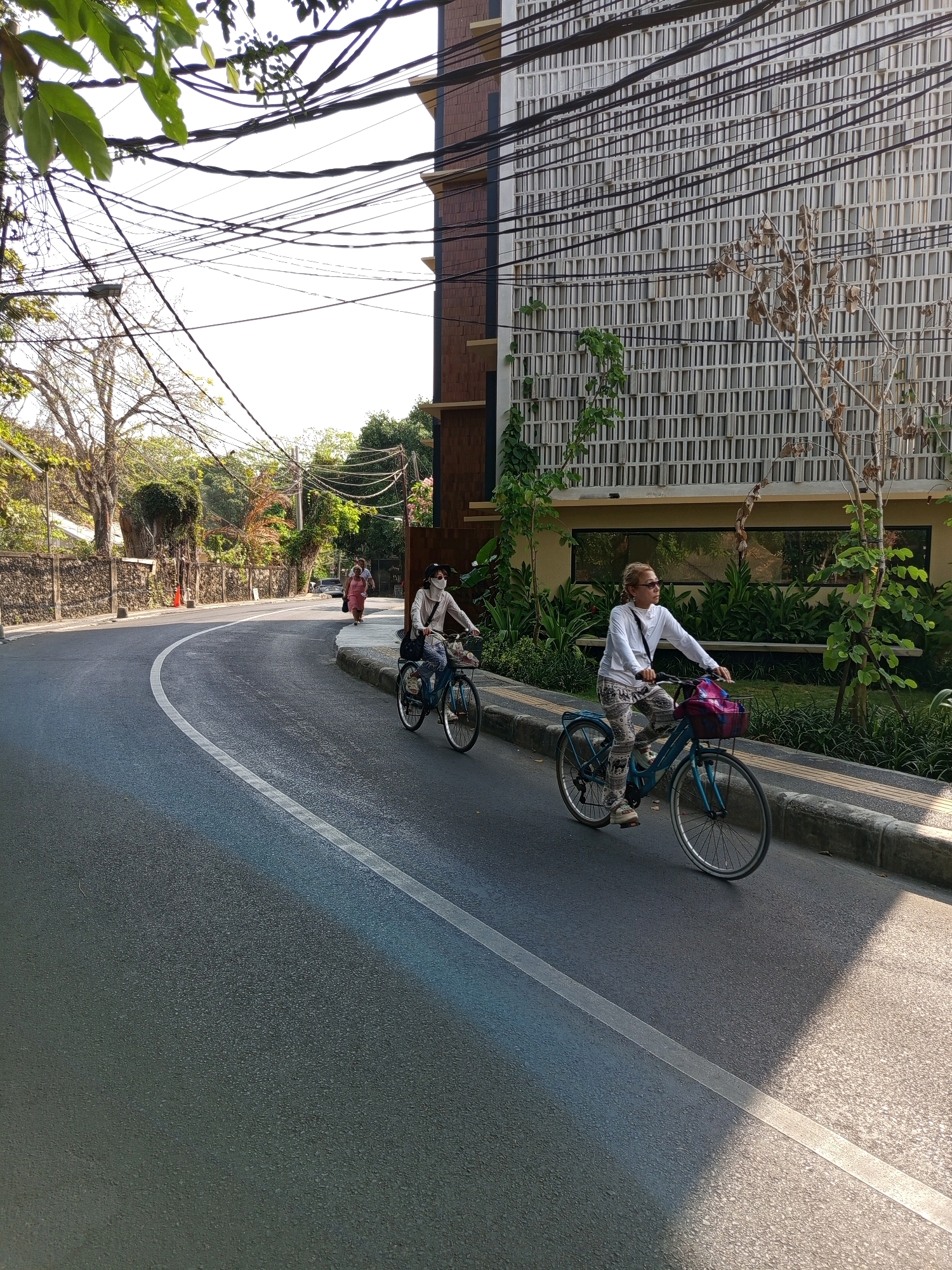 In Denpasar, Indonesia, on a paved street with a white lane marker, two individuals are cycling. In the foreground, a person with blonde hair wearing sunglasses, a white long-sleeved shirt, and patterned pants rides a light blue bicycle with a pink basket. They are on the right side of the road. Behind them and further down the street, another person wearing a white mask and a light-colored shirt rides a bicycle. They are positioned on the left side of the road, approaching the first cyclist. Further in the distance, a person in a pink dress walks on the sidewalk. The street is lined with a stone wall and vegetation on the left, and a modern building with a patterned facade and numerous electrical wires overhead on the right. The lighting suggests it is daytime, with sunlight casting shadows.