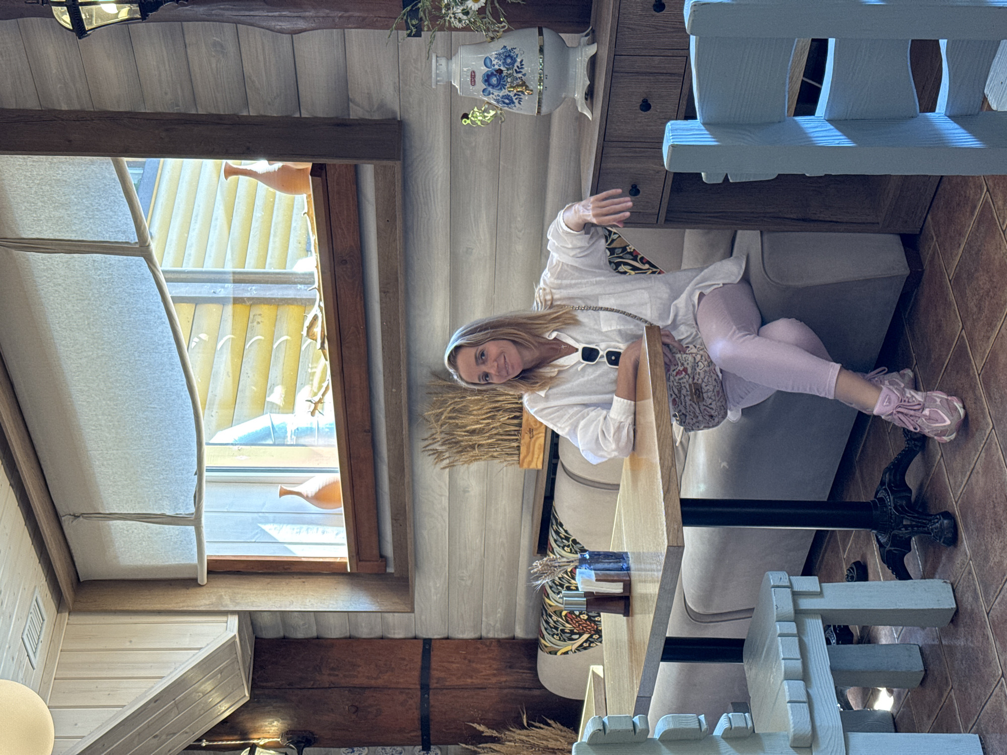 The image depicts a woman seated at a table in a cafe. The cafe has a rustic interior with wooden walls and a sloped wooden ceiling. A large window with slatted blinds reveals an outdoor scene. On the windowsill are two decorative vases. The woman is wearing a white shirt, light pink pants, and pink sneakers. She has a handbag slung over her shoulder. The table in front of her has salt and pepper shakers and a small vase with wheat stalks. Behind her is a banquette seating area with a patterned fabric. To the right of the banquette is a wooden cabinet topped with a decorative samovar and a small bouquet of flowers. The floor is tiled. The scene appears to be in Veliky Novgorod, Russia, suggested by the traditional decor elements such as the samovar and the rustic architecture.
