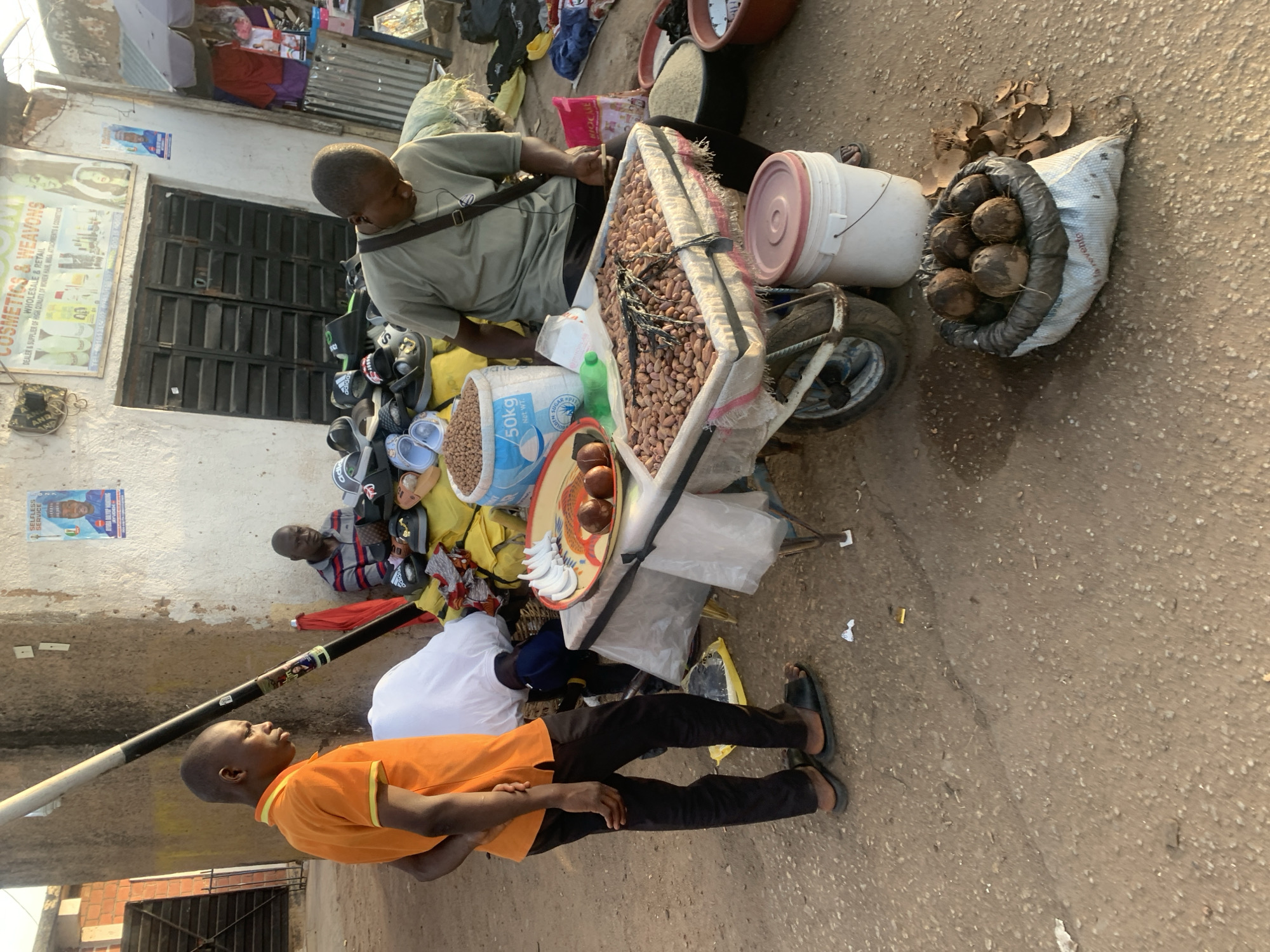 The image captures a street scene in Jos, Nigeria. A man sits next to a wheelbarrow filled with goods for sale, including peanuts in a large bag, coconuts in a sack, and other items. Another man, wearing an orange shirt and black pants, stands nearby. The street is paved, and there are buildings in the background with visible signage, including a cosmetics and weaving shop. Shoes are seen on the ground, and a poster is affixed to the wall. The scene is bathed in sunlight.
