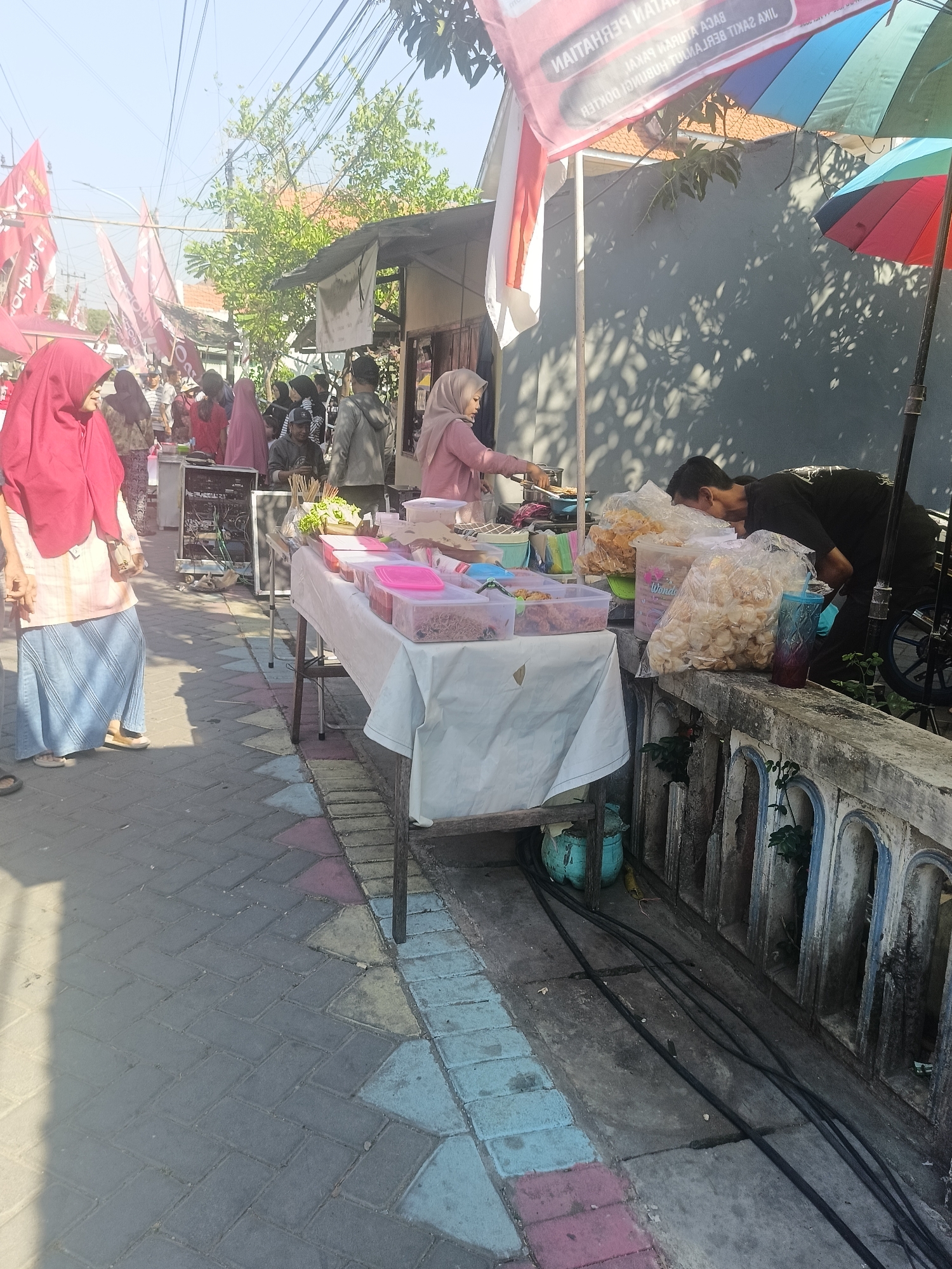This is a street-level, full-shot image taken in the city of Gedangan, Indonesia. The scene depicts a bustling outdoor market or gathering, with several people and various stalls. In the foreground, a woman wearing a red hijab and a blue skirt is standing on a cobblestone path. To her right, a table is laden with food items in plastic containers and bags of snacks, covered with a white tablecloth. Behind the table, two men are attending to the merchandise.
Further back, several other individuals are visible, some wearing hijabs. Red flags with white lettering are prominent in the background, suggesting a festive or political event. Overhead, power lines and trees are visible against a clear sky. A portion of a building and a colorful umbrella are also part of the scene, indicating the urban environment of the market. The ground is paved with interlocking bricks of various colors. The overall atmosphere appears to be active and commercial.