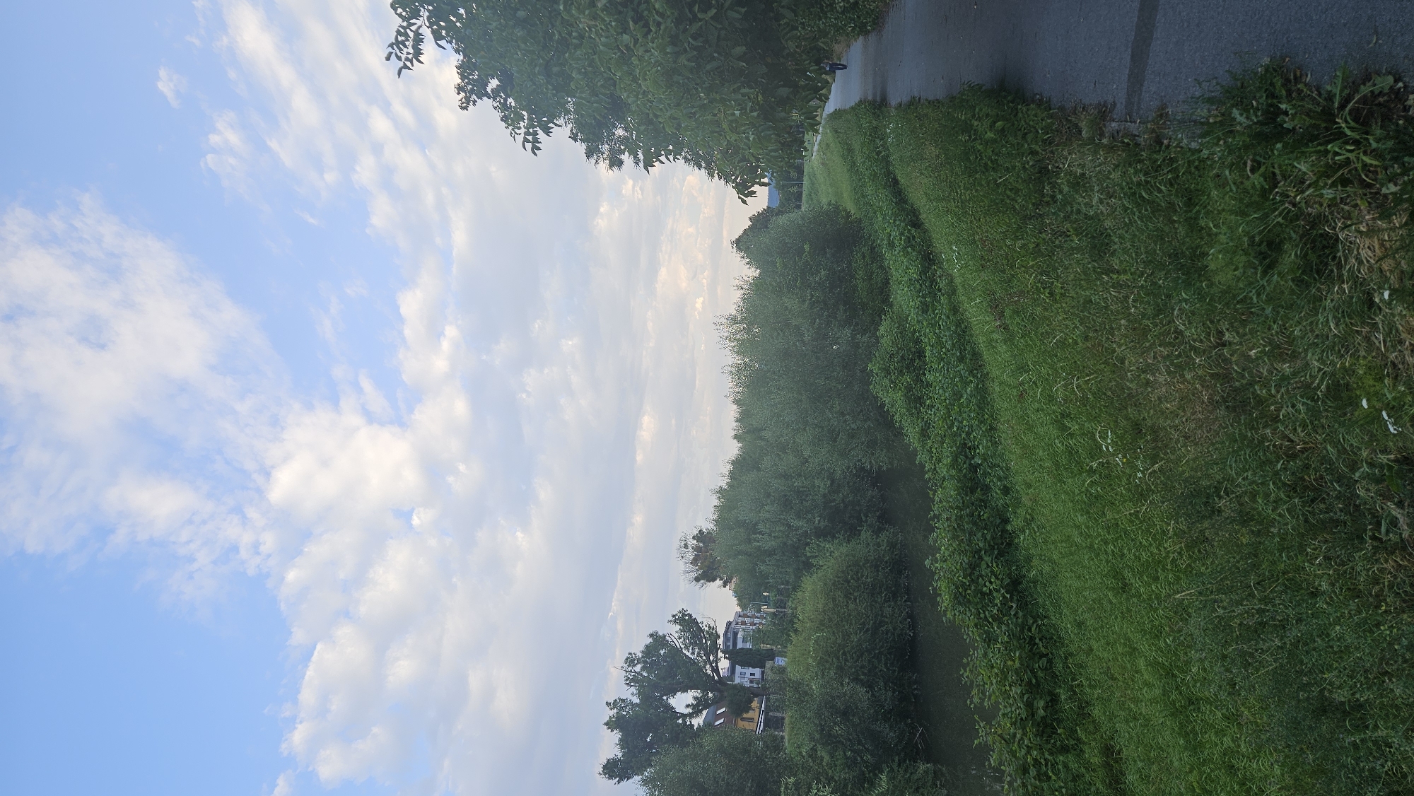 This is a vertical, eye-level shot of a paved pathway next to a river, with dense green foliage lining the banks and buildings visible in the distance on the left. The sky is a clear blue with scattered white clouds. In the far background on the right, a person is walking away from the camera on the pathway. The pathway appears to be a recreational path, possibly for walking or cycling. The river is calm and dark, reflecting the greenery on its banks. The overall scene depicts a peaceful, natural environment within an urban setting. The location is the Hornád riverbank in Prešov, Slovakia, a popular area for recreation and nature walks.