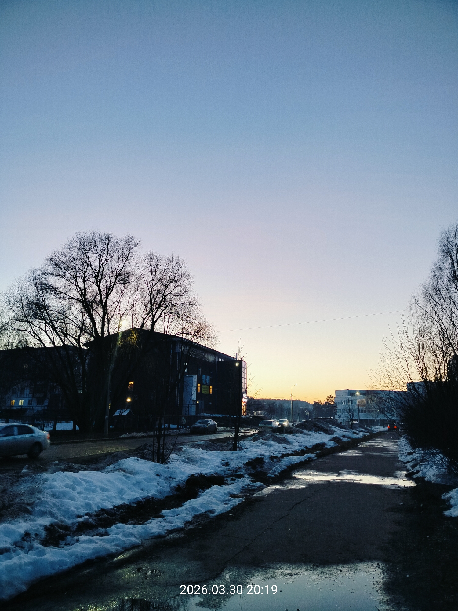 The image shows a street scene in Chaikovsky, Russia, on March 30, 2026, at 20:19. The sky transitions from blue to a lighter, sunset-tinged horizon. Buildings line the street, with bare trees silhouetted against the sky. Patches of snow remain on the sides of the road, which is wet and reflecting the sky. Cars are visible on the road. The lighting suggests twilight.