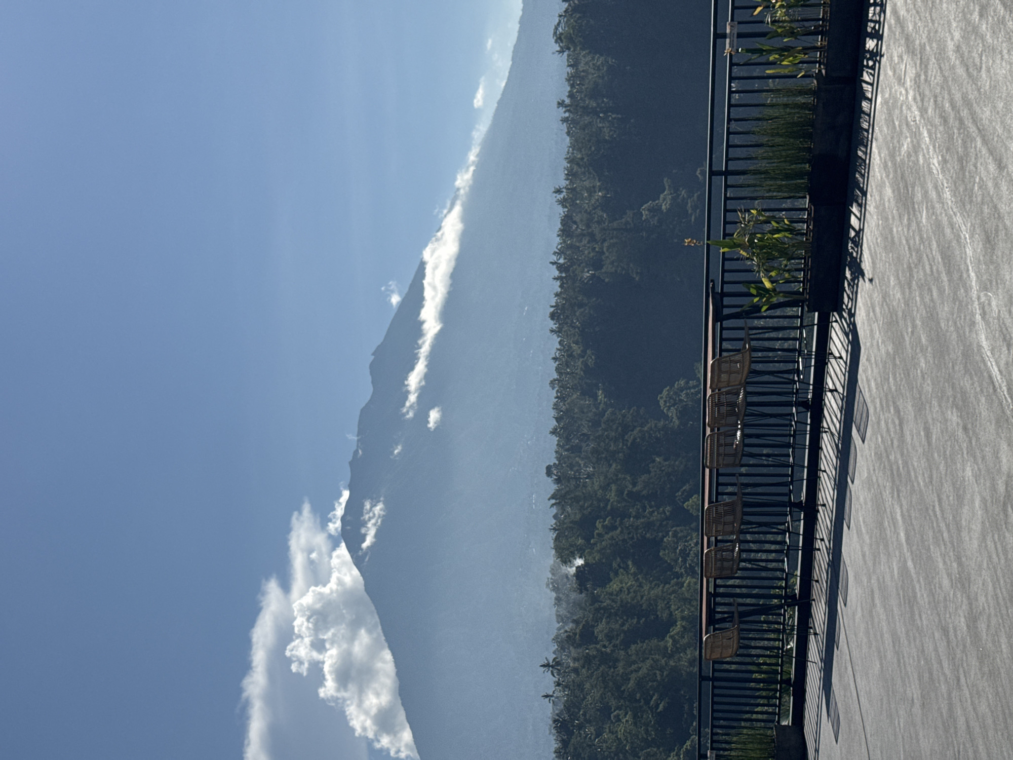 The image depicts a mountain landscape in Rendang, Indonesia, under a blue sky with scattered white clouds. To the left is a large mountain range, partially obscured by haze. The right side features dense green forest. A section of a building with a gray, textured surface is visible on the right, along with a black railing, potted plants, and several wicker chairs. The image is vertically oriented.
