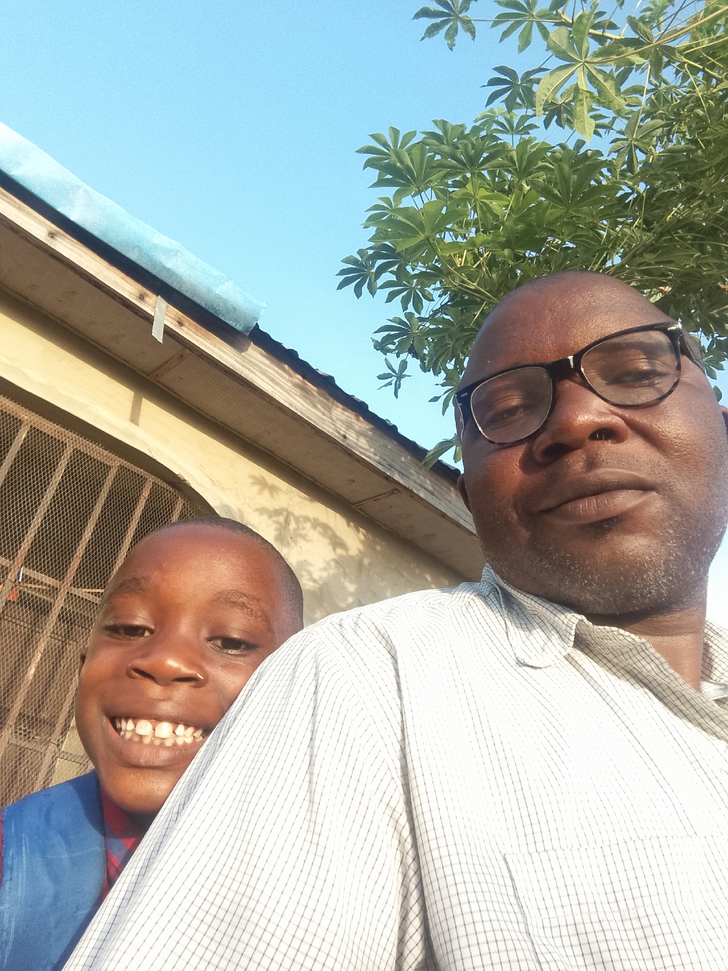 A selfie shows two dark-skinned people standing outside on a sunny day. A smiling child with gaps in his teeth is on the left. He wears a blue vest over a red shirt. A man with glasses and a graying goatee is on the right. He wears a light-colored, checked button-up shirt. Behind them is a building with a barred window and a section of roof that has blue material on top. A tree with large leaves is in the upper right corner, against a clear blue sky. The location is Unknown, Unknown.