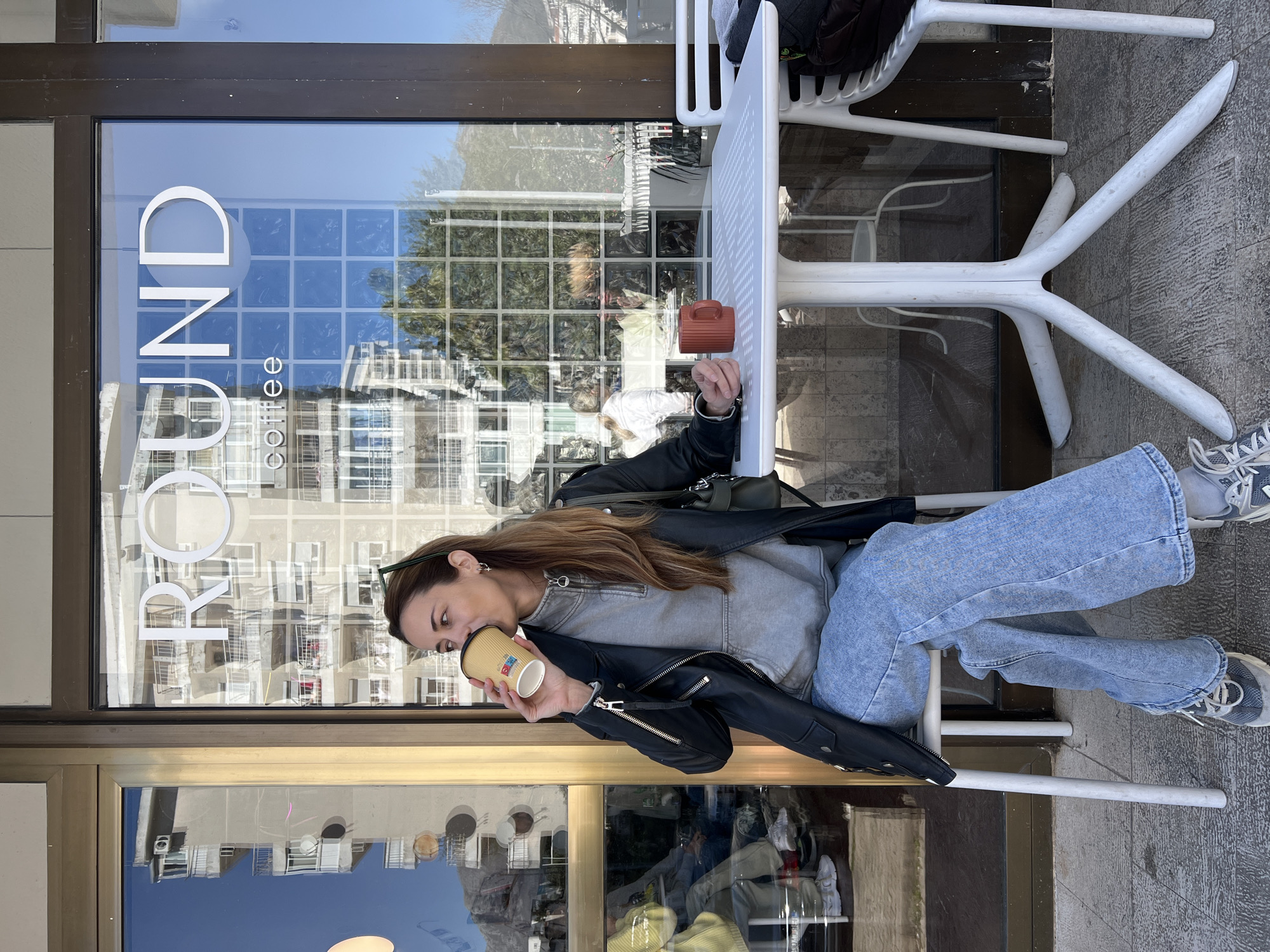 A woman sits at a white table outside a coffee shop in Bar, Montenegro. She drinks from a paper cup, dressed in blue jeans, a gray sweatshirt, and a black leather jacket. The sign on the shop window reads "ROUND coffee". A mug rests on the table next to her. The exterior of the shop reflects the opposite building, indicating an urban setting. White plastic chairs are at the table. The ground is tiled.