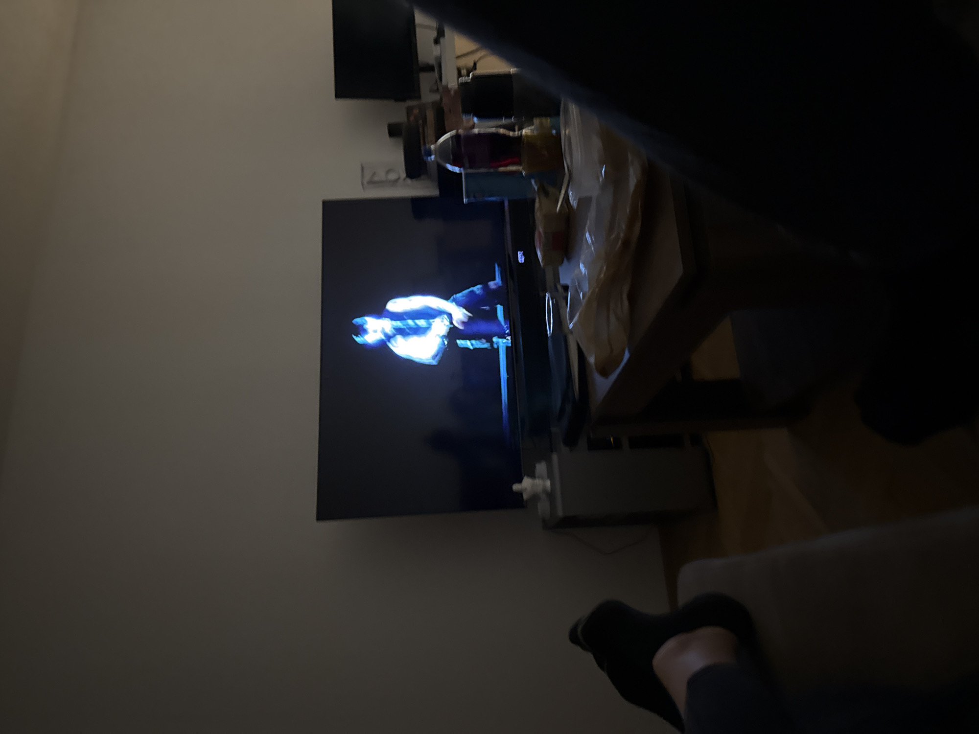 A person wearing black socks is sitting on a gray couch in a dimly lit room watching television. The television is displaying a scene of a male figure wearing a light blue jacket and dark pants. A gray table is located next to the television, with assorted items on top, partially obscured. In the background, a wall is visible. There is nothing in the image that indicates the location is Lovech, Bulgaria.