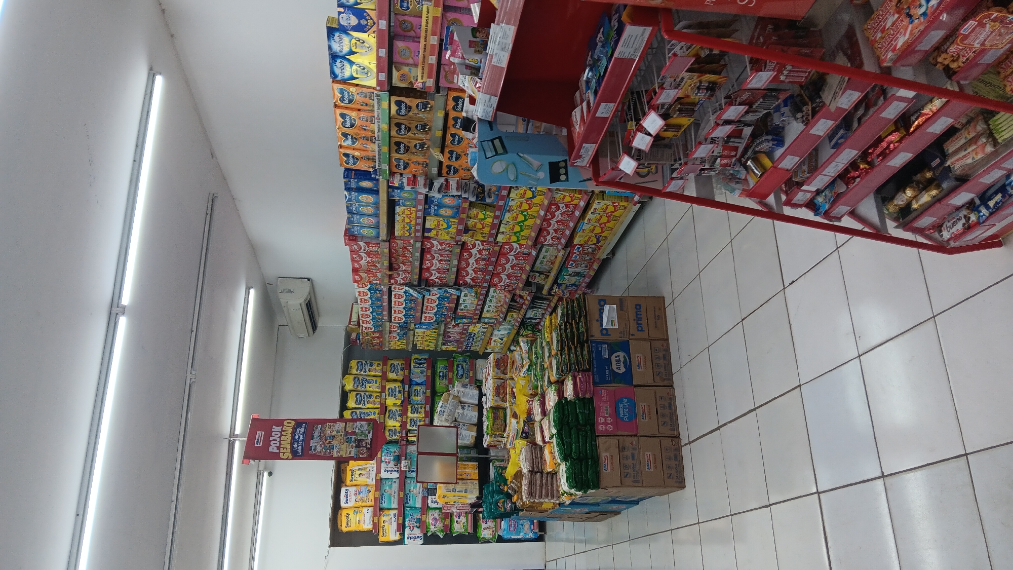 The image shows the interior of a convenience store, likely an Alfamart in Purwakarta, Indonesia. The store is well-lit with fluorescent lights on the ceiling. Aisles are stocked with various products, primarily baby formula, snacks, and beverages. In the foreground, there is a display rack with candy bars and other packaged goods. In the background, shelves are filled with boxes of baby formula and diapers. A sign hanging from the ceiling reads "POJOK SEMBAKO" which translates to "Grocery Corner" in Indonesian, indicating a section dedicated to essential groceries. An air conditioning unit is visible on the wall above some of the shelves. The floor is tiled with white square tiles. The scene is devoid of any people.
