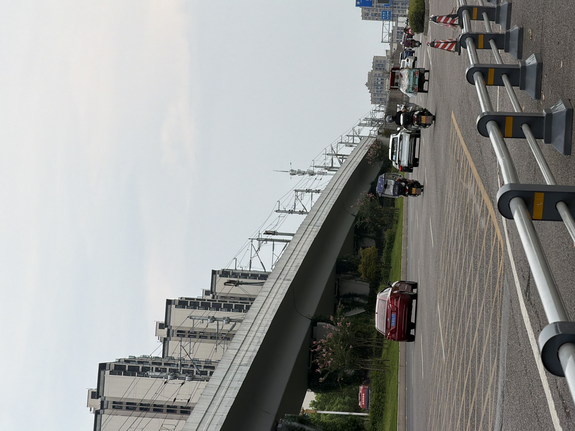 The image shows a street scene in Jiangjin, China, with a prominent elevated train track running diagonally across the frame. The train track is supported by concrete pillars and lined with overhead electrical wires. Several modern, multi-story apartment buildings are visible behind the train track, characterized by their light-colored facades and numerous windows.
The street itself is paved and appears to be a multi-lane road. Traffic includes several vehicles: a red sedan in the foreground, a white SUV, a small truck, and multiple motorcycles. People on motorcycles are dressed in casual clothing, with some wearing helmets. One motorcycle has a blue and white canopy over the passenger seat. The atmosphere appears overcast, with a pale, diffused light.
On the right side of the road, there are traffic cones and a metal railing. The road markings include yellow and white lines. Vegetation, including trees and bushes, is present along the sides of the road and under the elevated track. In the distance, more buildings and traffic are visible.