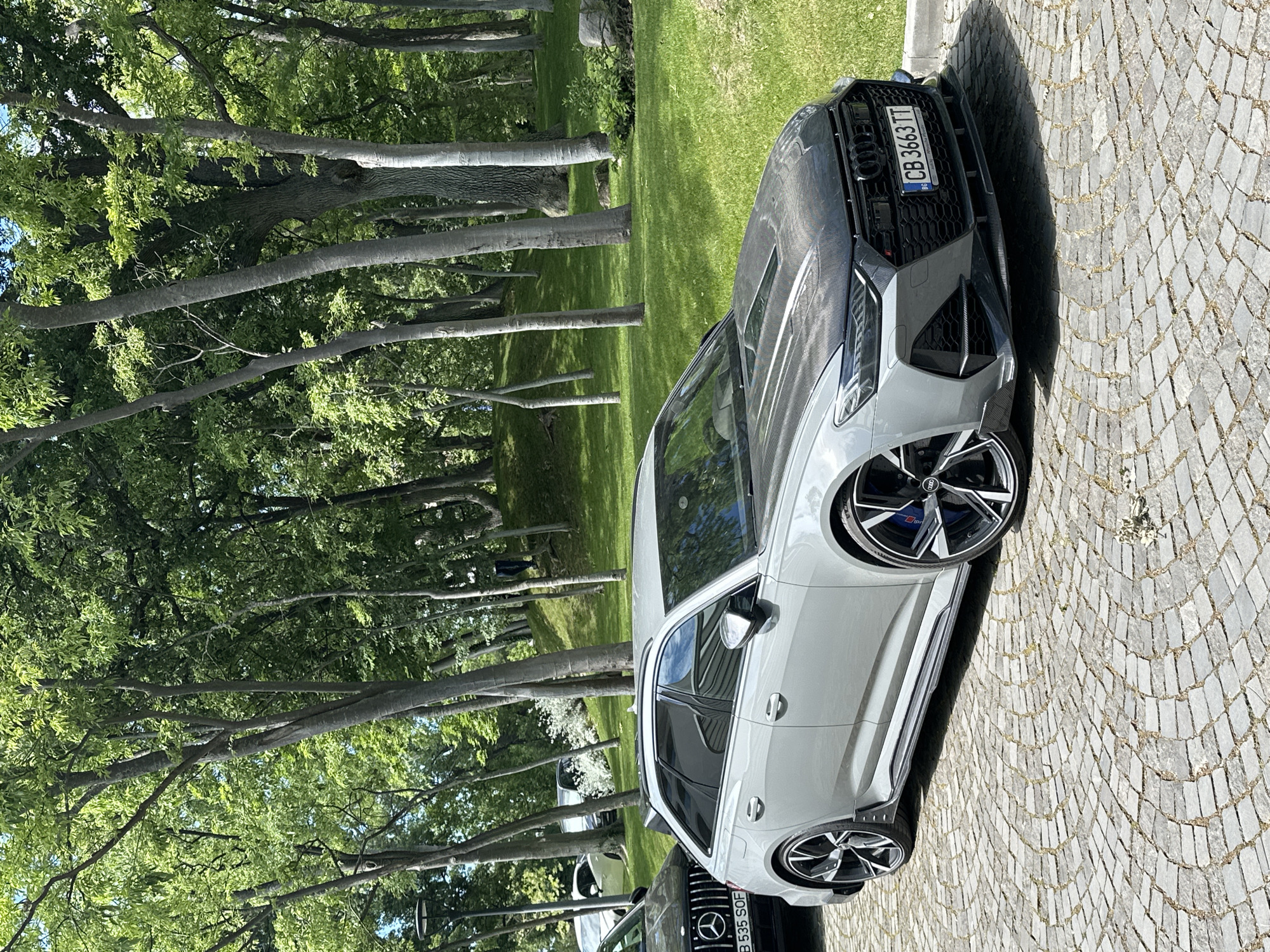 The image shows a light gray Audi RS6 Avant Performance parked on a cobblestone area near a park in Varna, Bulgaria. The car has a carbon fiber hood and black wheels. The license plate number is CB 3663 TT. Another car, a black Mercedes-Benz, is partially visible behind the Audi, with its license plate B 535 SOF. In the background, there is a grassy area and a row of tall trees providing shade, possibly a park or garden. The lighting suggests it is a sunny day. There are no people visible in the immediate vicinity of the cars.