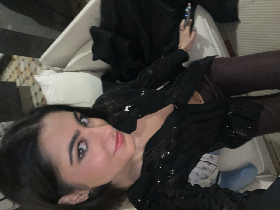 A woman with dark hair and red lipstick lies on a white couch, wearing a sparkly black shirt and brown pants. She looks directly at the camera. In her right hand, she holds a cell phone. A dark jacket rests behind her. The couch is covered with white and patterned pillows, with a glimpse of a tiled floor in the background. The image appears to have been taken indoors with casual lighting.