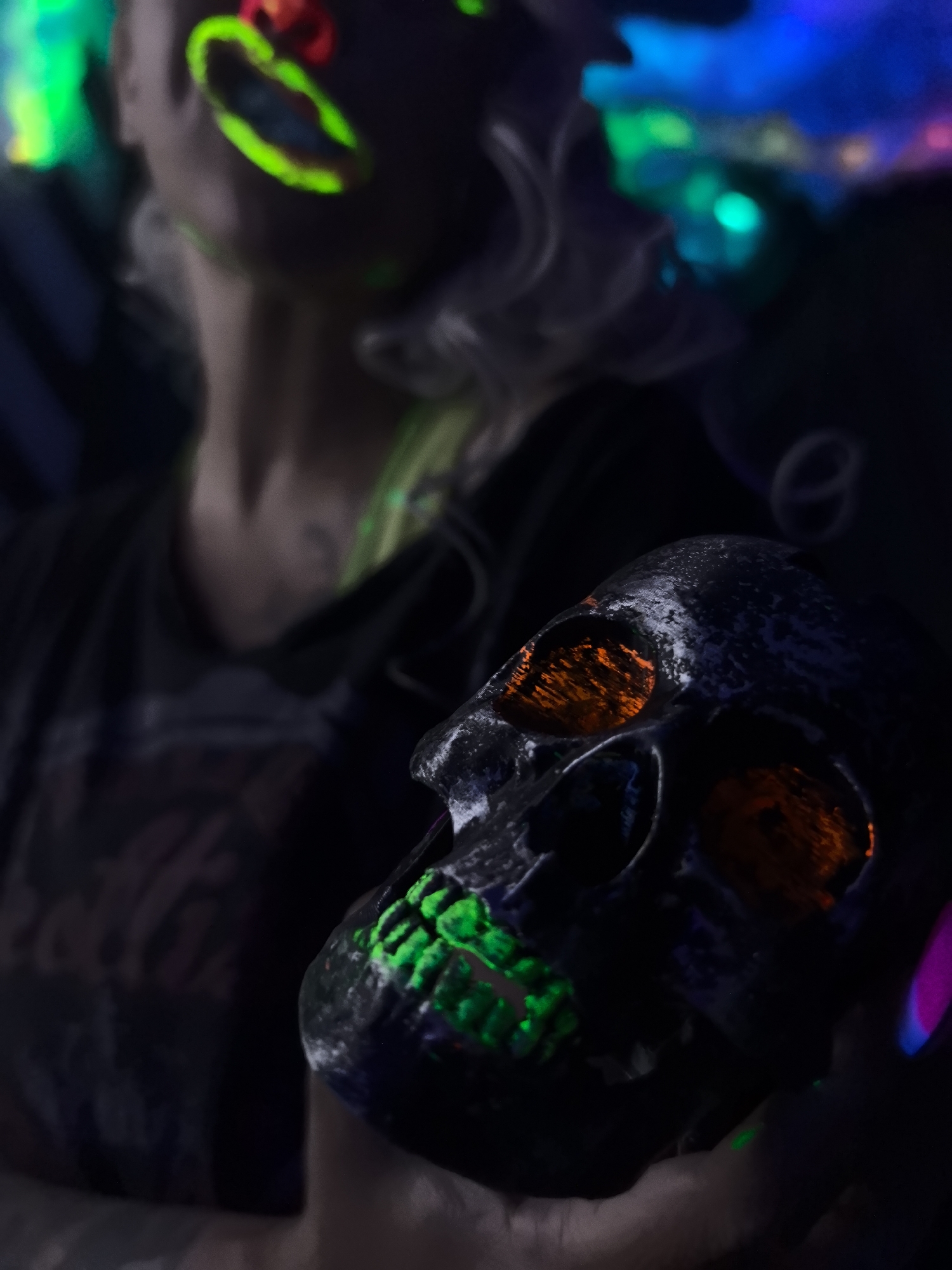 The image depicts a close-up of a person holding a skull with glowing neon paint on it. The skull has orange-glowing eye sockets and green glowing paint on its teeth and jaw. The person's face is partially visible in the background, also adorned with neon green and red paint on their lips and around their eyes. The overall lighting is dark, with vibrant neon colors illuminating parts of the scene, suggesting a setting with blacklights. The background shows blurry lights in shades of blue and green, and hints of white smoke or mist. There are no discernible architectural or environmental details that would place this scene in a specific location, such as Noviy, Russia. The focus is on the dramatic lighting and the Halloween-themed skull.