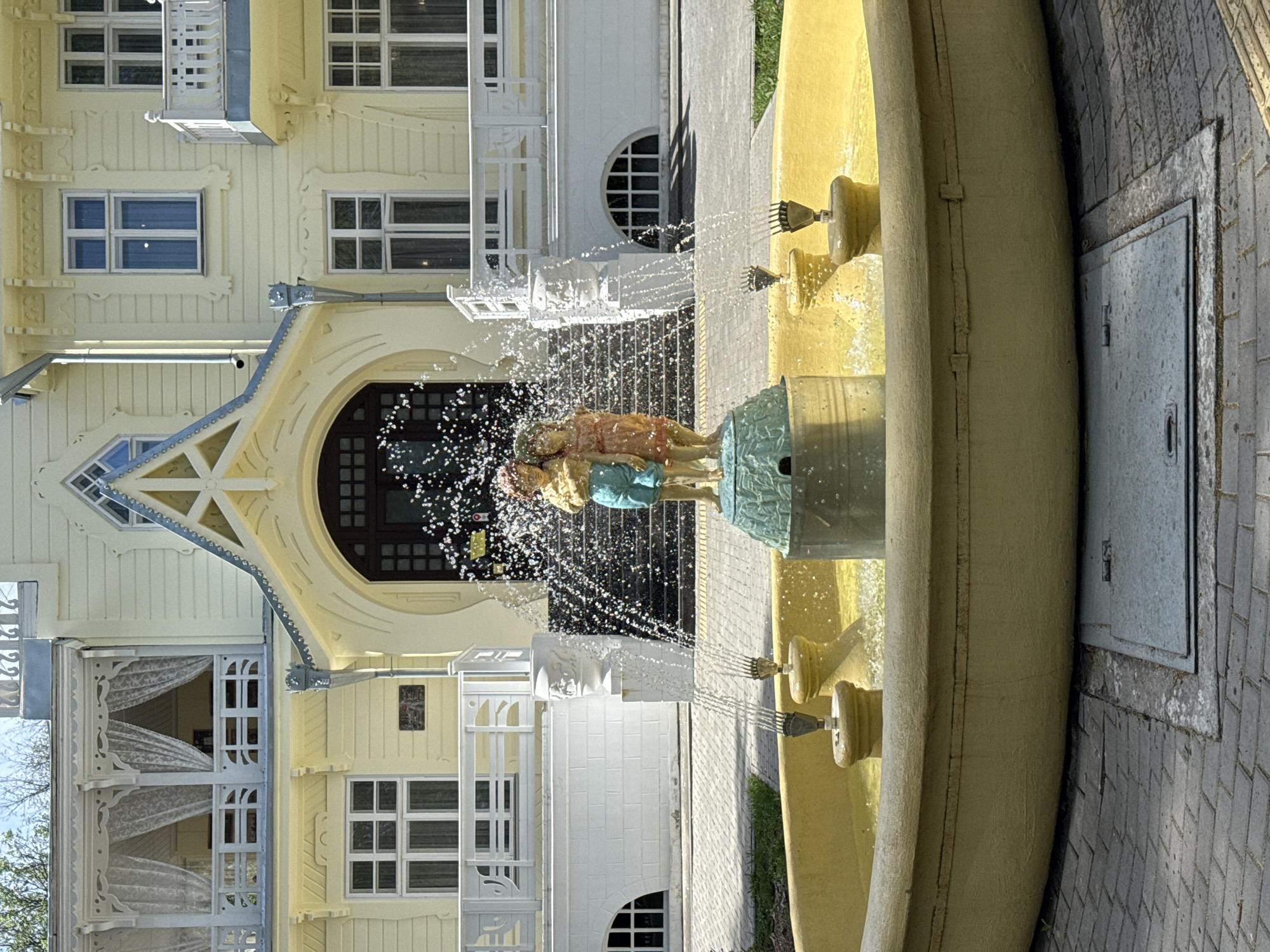 The image depicts a fountain in the city of Sarapul, Russia, situated in front of a yellow building with ornate architectural details and white accents. The fountain is circular with a yellow exterior, and it features a sculpture of figures at its center. Water is actively spraying from various points on the fountain, creating a visual effect of motion and sparkle. The building behind the fountain has multiple windows and a distinctive triangular gable over the entrance. A section of brick paving is visible in the foreground near the fountain.