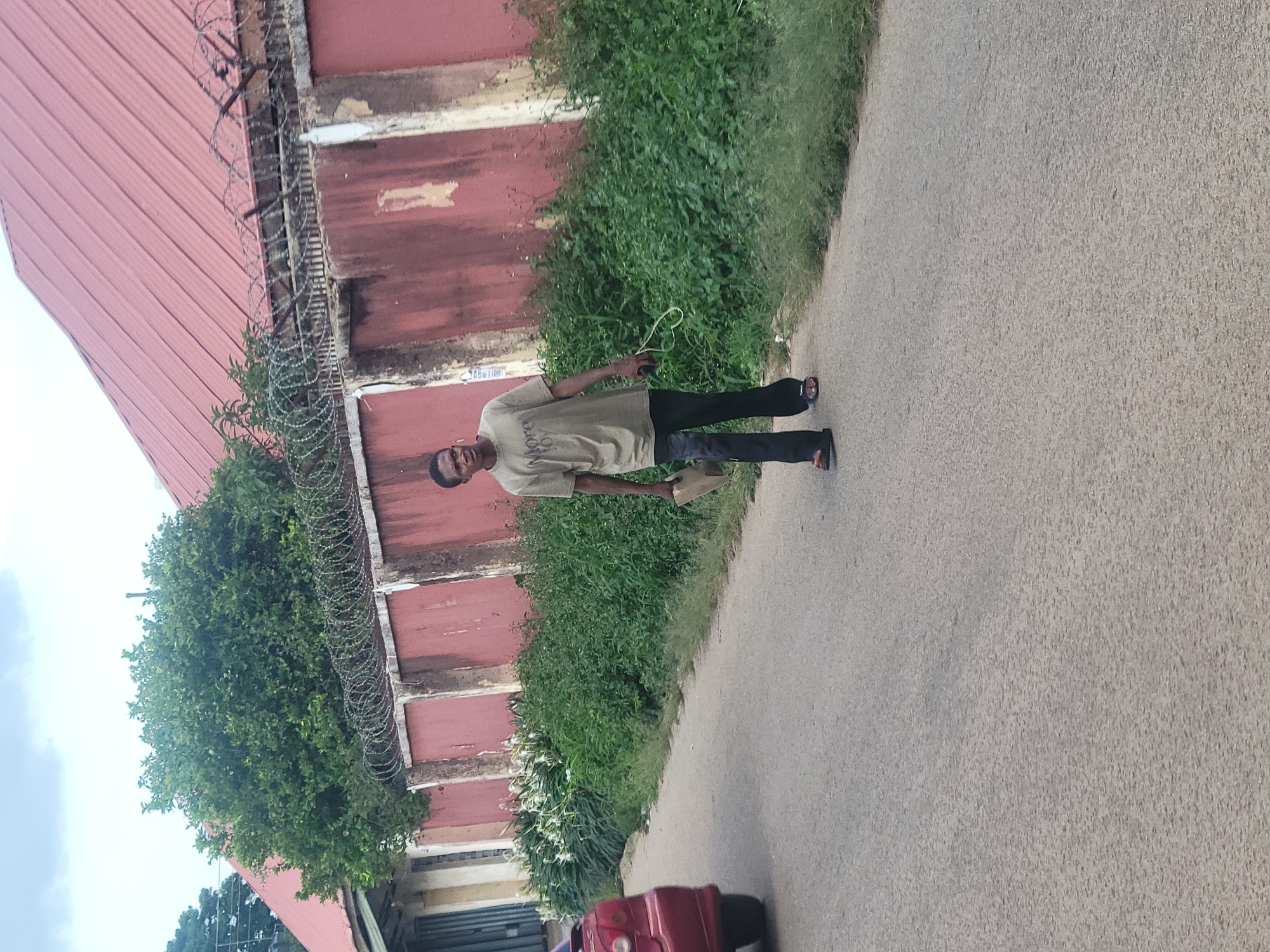 A young man walks along a paved road in Jos, Nigeria, wearing a light brown t-shirt and dark pants, with his right hand holding a light-colored bag or book, and his left hand holding a pair of white earphones. He is looking towards the viewer, with his mouth slightly open. The road is flanked by lush green vegetation, and behind the vegetation is a red-colored wall topped with barbed wire. A red car is partially visible on the left side of the road, moving away from the camera. The sky above is overcast.
