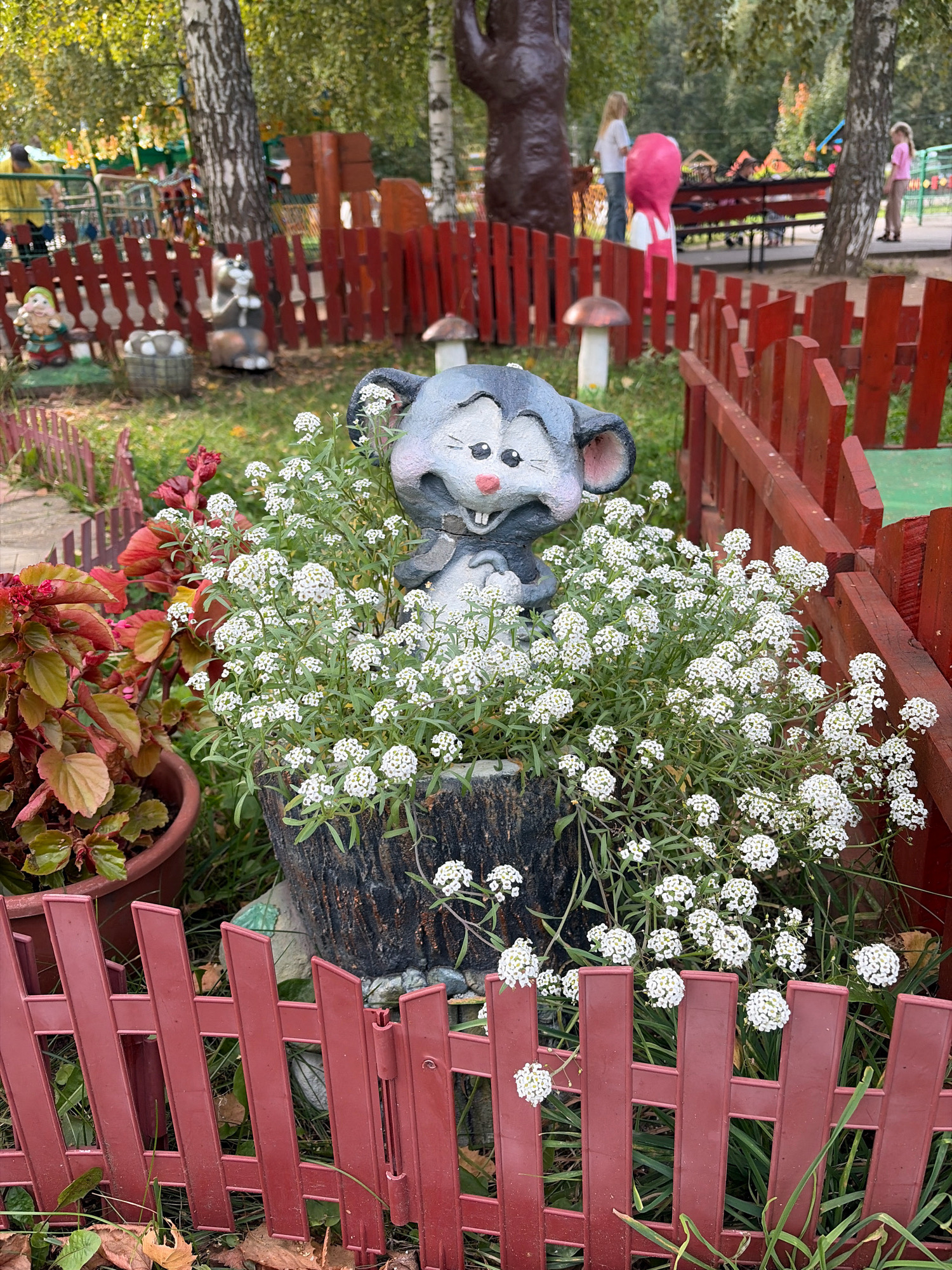 This image depicts a decorative garden scene in Chaikovsky, Russia, featuring a ceramic mouse statue nestled amongst white flowers. The mouse, with its ears perked and a slight grin, appears to be peeking out from a cluster of small white blooms. The statue is placed in a dark, textured planter that resembles a hollowed-out log. To the left of the mouse, a potted plant with large, reddish-brown leaves adds a splash of contrasting color. The entire arrangement is framed by a low, red wooden fence with a decorative picket design. In the background, a playground area is visible with children and playground equipment, suggesting a public park or a family-oriented recreational space within the city. The trees in the background indicate an outdoor setting with deciduous foliage.
