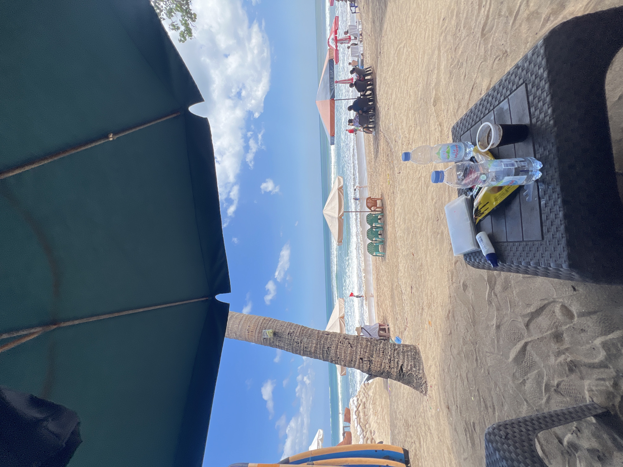 The image depicts a beach scene in Kuta, Indonesia, on a bright, sunny day. In the foreground, a dark, woven table holds two bottles of water and a cup of dark liquid. Behind the table, a palm tree stands prominently, its trunk weathered and textured. The beach itself is sandy, with a few footprints visible. In the middle ground, beach umbrellas provide shade for a group of people sitting at tables. Further back, the ocean stretches to the horizon under a bright blue sky dotted with white clouds. Waves crash onto the shore, and a few individuals can be seen swimming or walking near the water. Surfboards are propped up on the left side of the image. The overall atmosphere is one of relaxation and leisure on a tropical beach.
