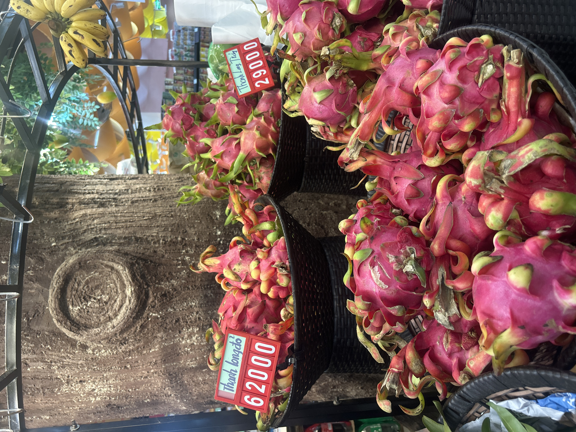 Bắc Nha Trang, Vietnam: Three black woven baskets hold red-skinned dragon fruits, with price signs reading "Thanh Long 29000" and "Thanh Long do 62000". Above, bananas hang from a metal rack. The background features a textured surface.