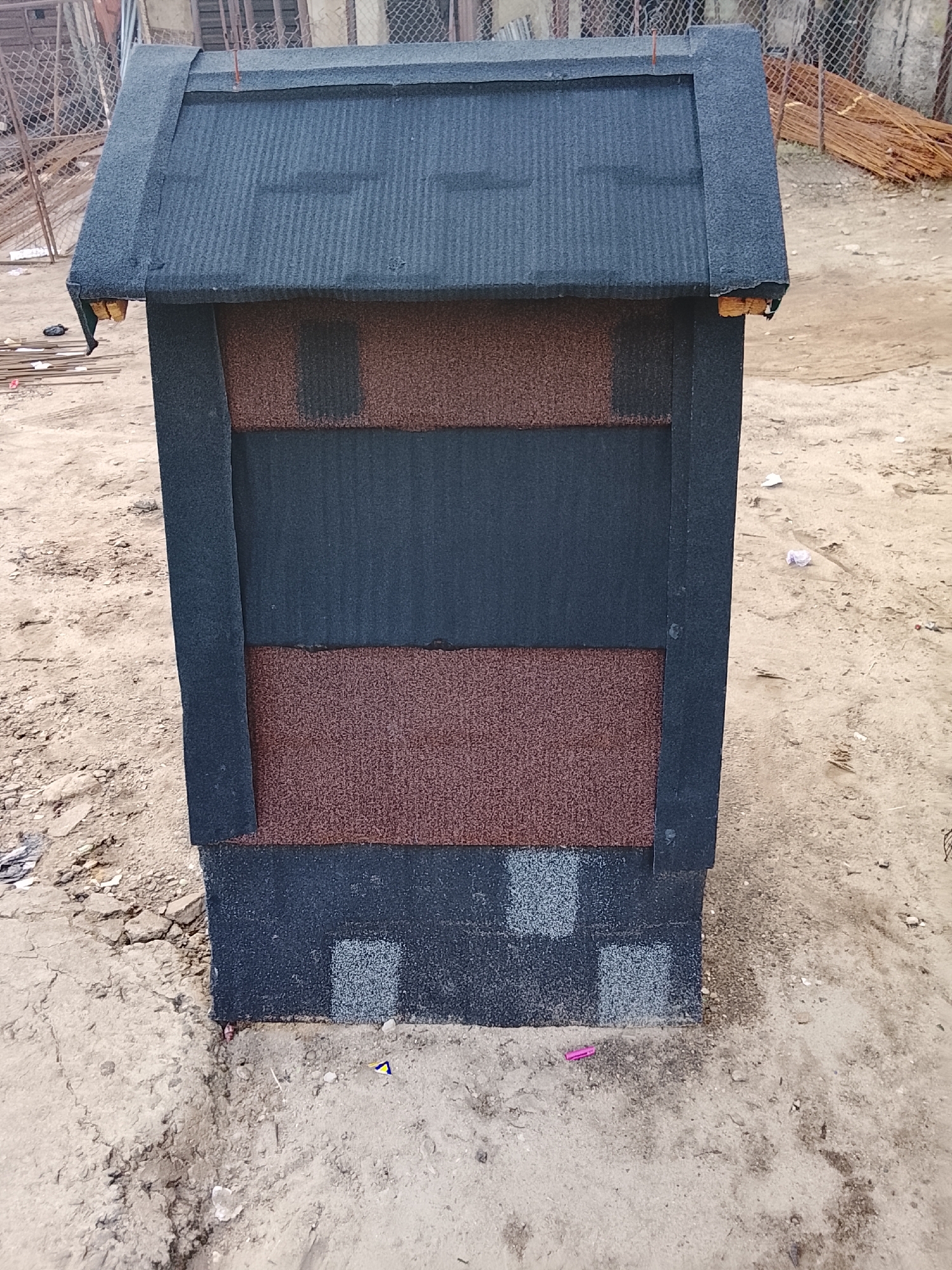 A dark gray, gable-roofed structure, likely a small house or shed, stands on a dirt ground in Rumuepirikom, Nigeria. The roof is covered with dark gray shingles. The walls of the structure are a combination of dark gray and reddish-brown panels, also made of shingles. The bottom portion of the walls features a pattern of square, light gray shingles. The background shows a chain-link fence, some rebar, and scattered debris. The ground is a mixture of sand, dirt, and small stones. There are no people visible in the image.