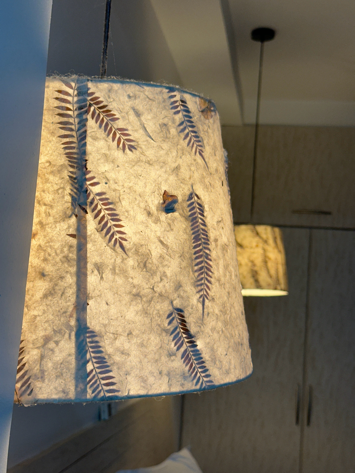 The image captures a close-up view of a hanging lampshade, illuminated from within, casting a warm glow. The lampshade is made of textured, translucent paper adorned with a pattern of dark, leaf-like silhouettes. A thin wire suspends the lampshade from a dark ceiling fixture. In the background, another out-of-focus lampshade with a similar patterned design is visible. The environment appears to be an indoor setting, with a neutral-colored wall and cabinetry visible in the blurred background. The overall impression is one of soft, ambient lighting within a domestic or commercial space. No specific location in Lalitpur, Nepal can be determined from the image itself.