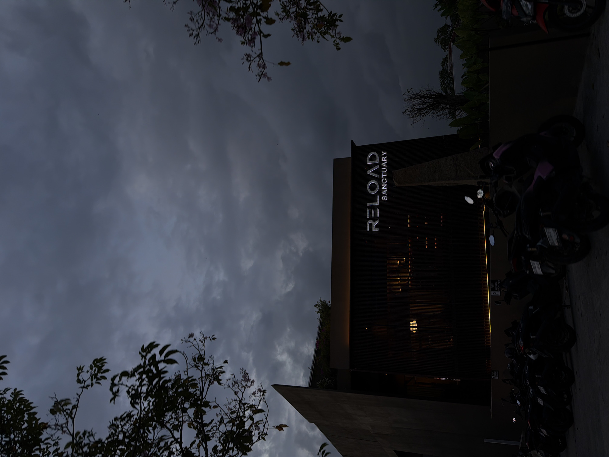 Captured at night, the image shows the exterior of "RELOAD SANCTUARY" in North Kuta, Indonesia. The building, identifiable by the illuminated signage, has a modern design. Parked motorcycles line the building's side on the right. Silhouetted trees and an overcast sky form the backdrop.