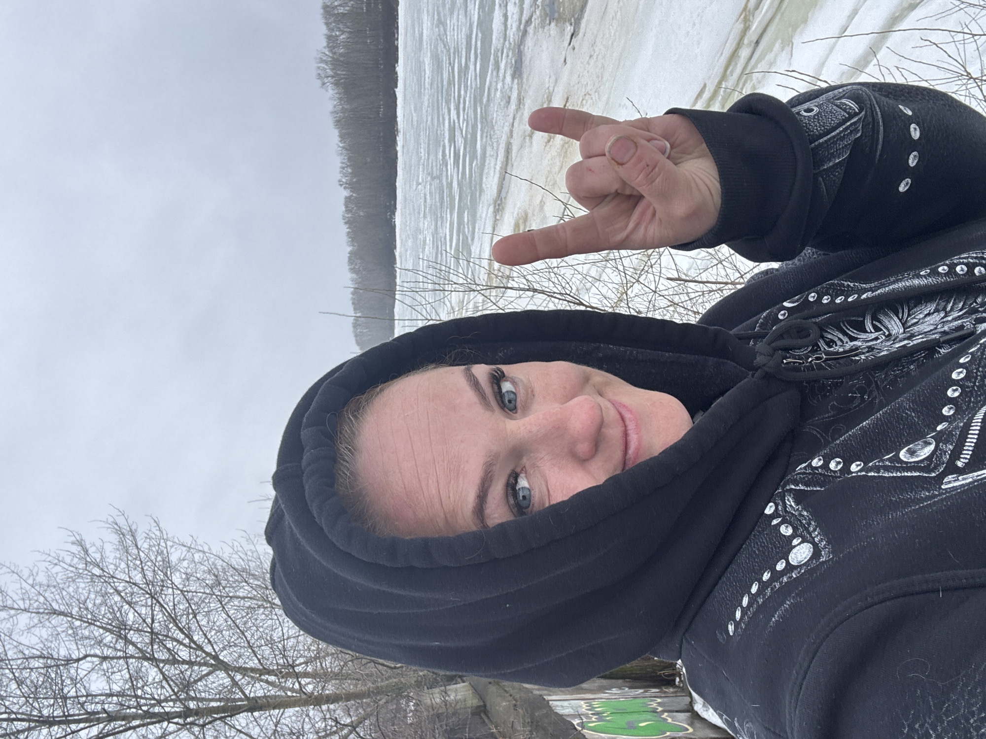 A woman with fair skin and blue eyes is wearing a black hooded sweatshirt and making a "rock on" hand gesture. She is positioned in front of a frozen river or lake. Bare trees are visible in the foreground and along the shoreline. The sky is overcast. The location is Chaikovsky, Russia, based on image metadata.