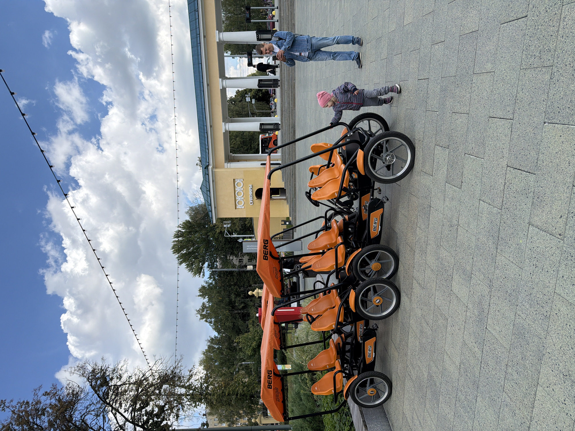 The image depicts an outdoor scene in Nizhny Novgorod, Russia, likely in a park or public square. In the foreground, a row of four orange pedal karts, branded with "BERG," are parked. These karts have canopies and multiple seats. A young child, wearing a pink hat and patterned jacket, stands near the karts, touching one of them. To the right, an adult male, dressed in a denim jacket and jeans, is holding a smartphone and appears to be taking a picture or video. In the background, there is a light yellow building with a green roof, supported by large white columns. The building has arched doorways and a sign that reads "СУВЕНИРЫ" (Souvenirs). Trees and a cloudy blue sky with some string lights overhead complete the scene.