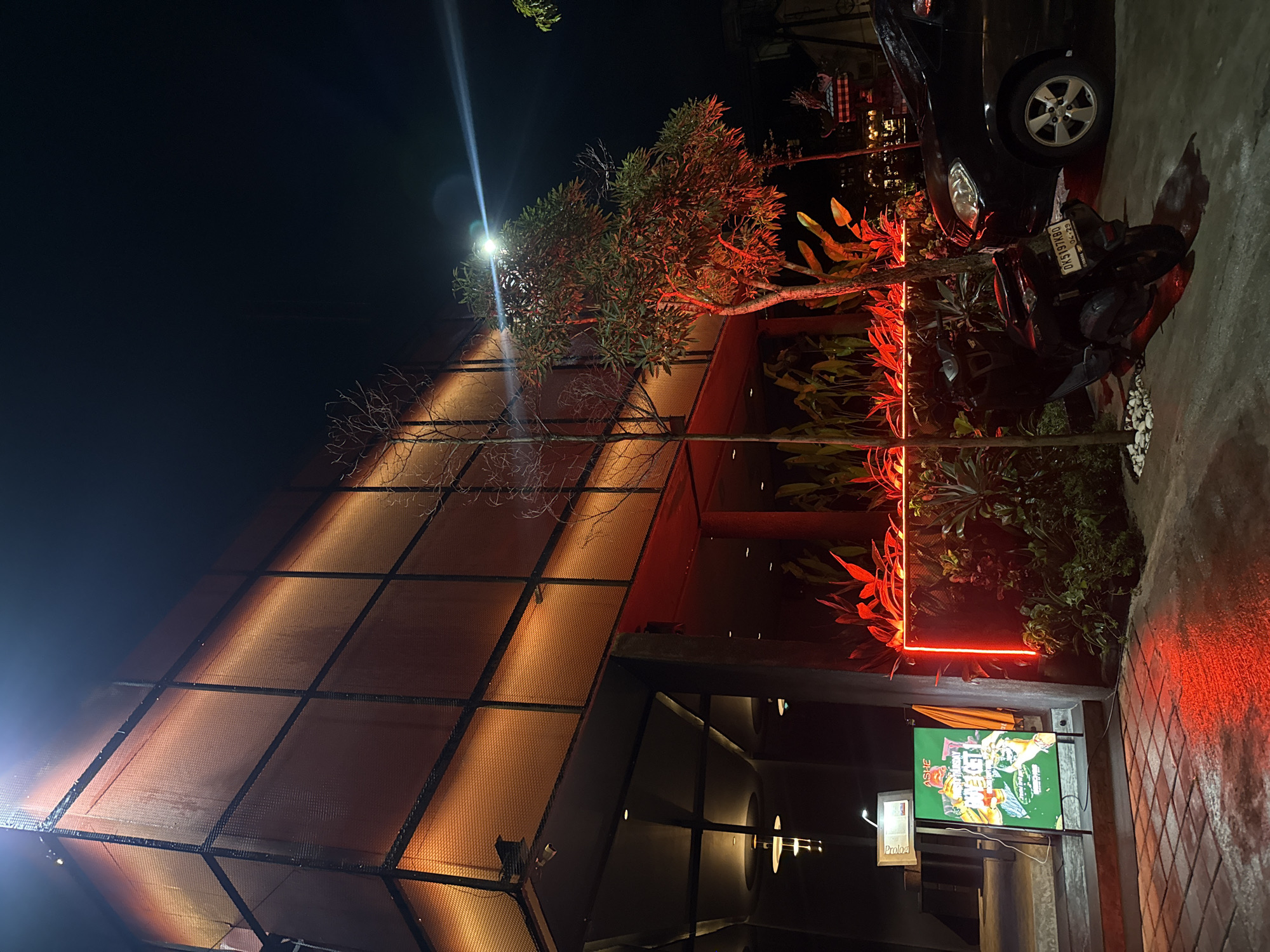 The image displays a modern-style building at night, likely a commercial establishment such as a restaurant or bar. The building features an angular, multi-faceted facade made of a textured, possibly mesh or perforated, material that glows with a warm, orange-yellow light from internal or external illumination. A bright white light source is visible high above, cutting through the dark sky and partially illuminating tree branches.
In front of the building, there are several trees and lush green plants. A line of red neon lights brightly illuminates some of these plants and outlines a section of the building's base, creating a striking contrast with the warm facade.
To the right, a black car and a dark-colored motorcycle are parked on a paved surface, which appears somewhat wet or reflective under the artificial lights. The motorcycle's license plate, "DK 5197 RBO" with an expiry date "09-23," is clearly visible, suggesting a location in Bali, Indonesia.
Near the entrance, an illuminated sign is present. The word "ASHE" can be seen on its top left corner. Below it, another sign has the word "Prologa" visible. The main content of the larger sign is a vibrant, graphic design with indistinct text and illustrations, possibly advertising menu items or events.
No people are visible, and no specific activity is taking place; it's a static scene captured at night. The atmosphere is one of contemporary urban nightlife, characterized by dramatic lighting and distinctive architecture.