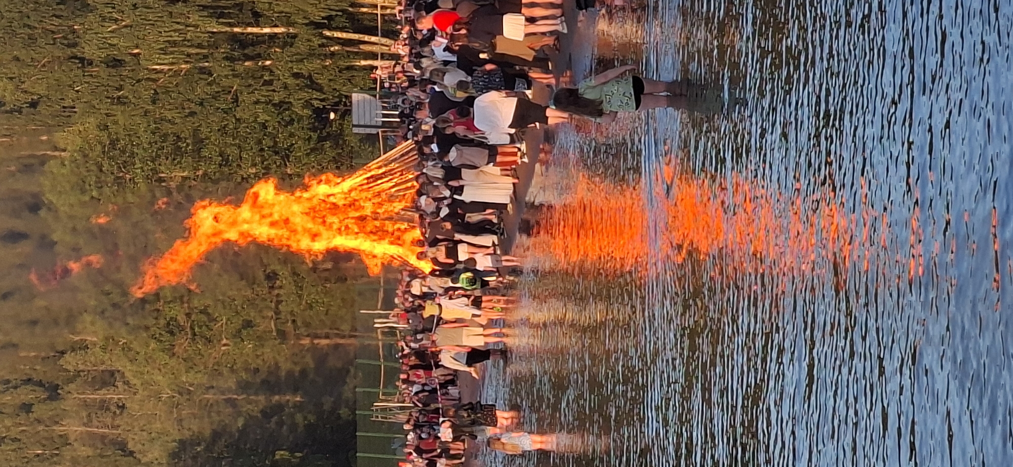 In Salpakangas, Finland, a large bonfire burns intensely, casting an orange reflection on the water in the foreground. A crowd of people are gathered around the bonfire, some standing in the water and others on the shore. The crowd appears to be observing the bonfire. The background consists of trees and a clear sky. The scene depicts an outdoor event with a significant fire and a large group of attendees.