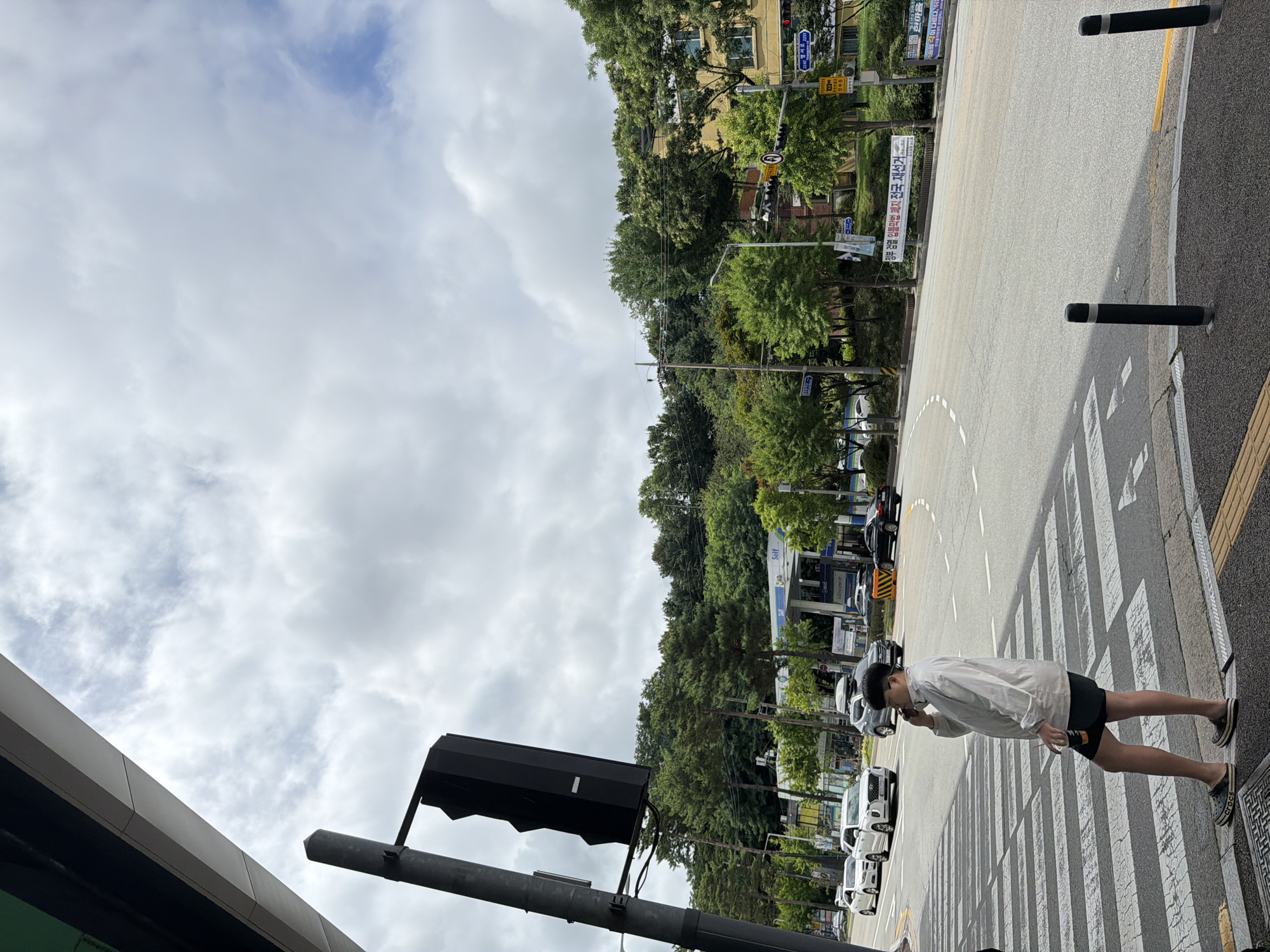 The image shows a street scene in Chuncheon-si, South Korea, under a cloudy sky. In the foreground, a pedestrian wearing a white jacket and black shorts is standing on a crosswalk, holding a cup and talking on a cell phone. To the left, a traffic light pole with a large black traffic signal is visible. The street is lined with trees and buildings, including a gas station with "Self" branding. Cars are present on the road, and traffic signs are visible. The overall environment suggests an urban setting on a somewhat overcast day.