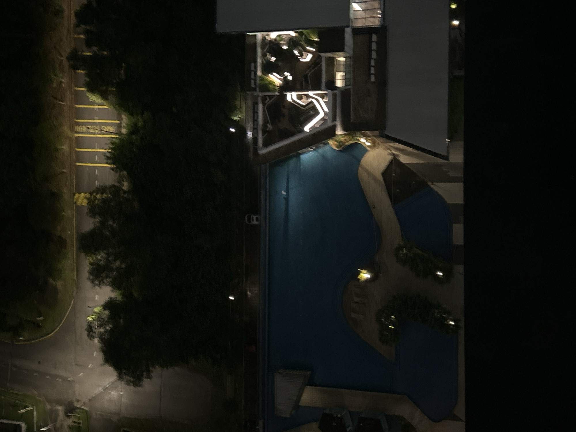 The image shows an aerial view at night, likely from a high-rise building, possibly in Kuala Lumpur, Malaysia. A swimming pool with an irregular shape is the central focus, illuminated by scattered lights. Adjacent to the pool is a modern building with white, illuminated walkways and structures on the roof. To the left of the pool and building is a dark green area of trees and vegetation. Further left is a road with yellow lines, presumably a parking area or loading zone. The scene is dark overall, with the artificial lights highlighting specific areas. No people are visible.