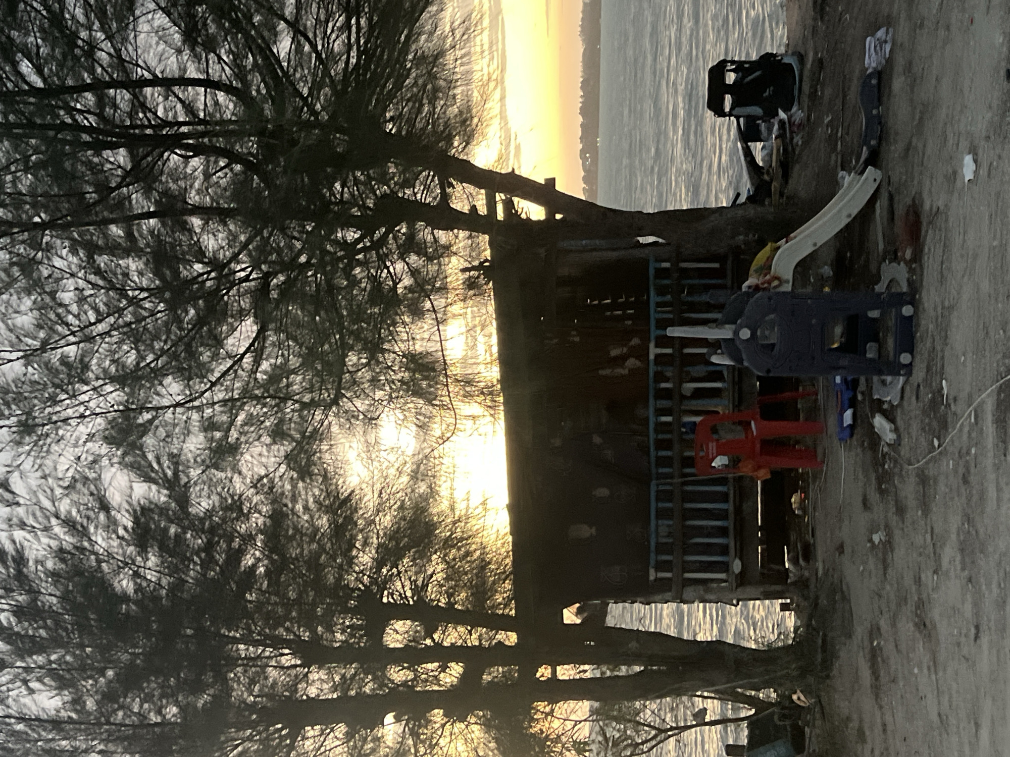 This is a medium-shot, eye-level, outdoor, candid photograph taken at dusk. In the foreground, a dilapidated wooden structure with a corrugated metal roof sits on a sandy, debris-strewn shore. A blue and white railing runs along the front of the structure. In front of the structure, a red plastic chair is positioned next to a dark blue and white plastic play structure with a white slide. To the right of the play structure, a black object that appears to be a stroller or a cart is visible. The ground is covered with scattered debris, including plastic bags and white paper. Two trees with thin trunks and dense foliage frame the scene on the left and right. The background features a body of water, likely the sea, with gentle waves. The sky is a gradient of pale yellow and grey, indicating a sunset. The overall environment suggests a neglected, coastal area, possibly in the Ko Pha-ngan District of Thailand. There are no people visible in the image.