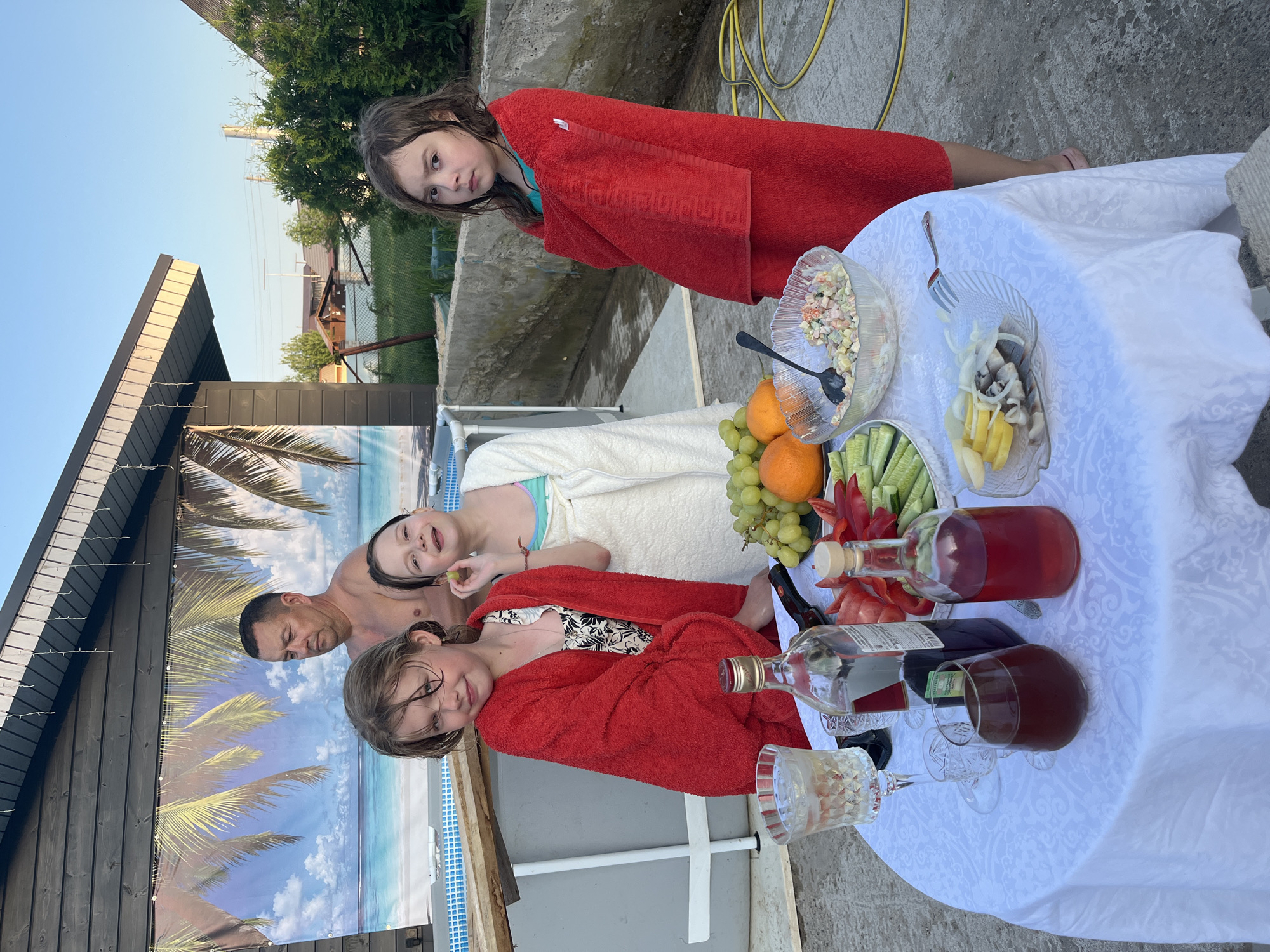 A group of people are gathered near a pool, likely for a meal after swimming. The scene is outdoors, possibly a backyard in Новая Чернушка, Russia. Three girls are present, two wearing red towels and one in a white towel. A man with no shirt is also present. A table covered with a white tablecloth holds a variety of food and drinks, including salad, fruit, vegetables, and bottles of liquid, possibly wine. There are also glasses. A backdrop with a beach scene is set up behind the pool.