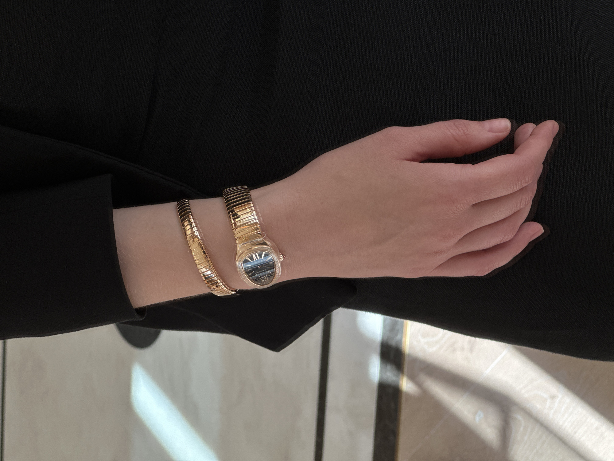 The image shows a close-up of a person's wrist wearing a rose gold watch with a serpentine bracelet. The watch has a dark dial with Roman numerals. The person is wearing a black garment, possibly a jacket or dress. The background appears to be an indoor setting with a tiled floor that has light and shadow patterns. No specific landmarks or features are visible to identify a location in Tel-Aviv, Israel.