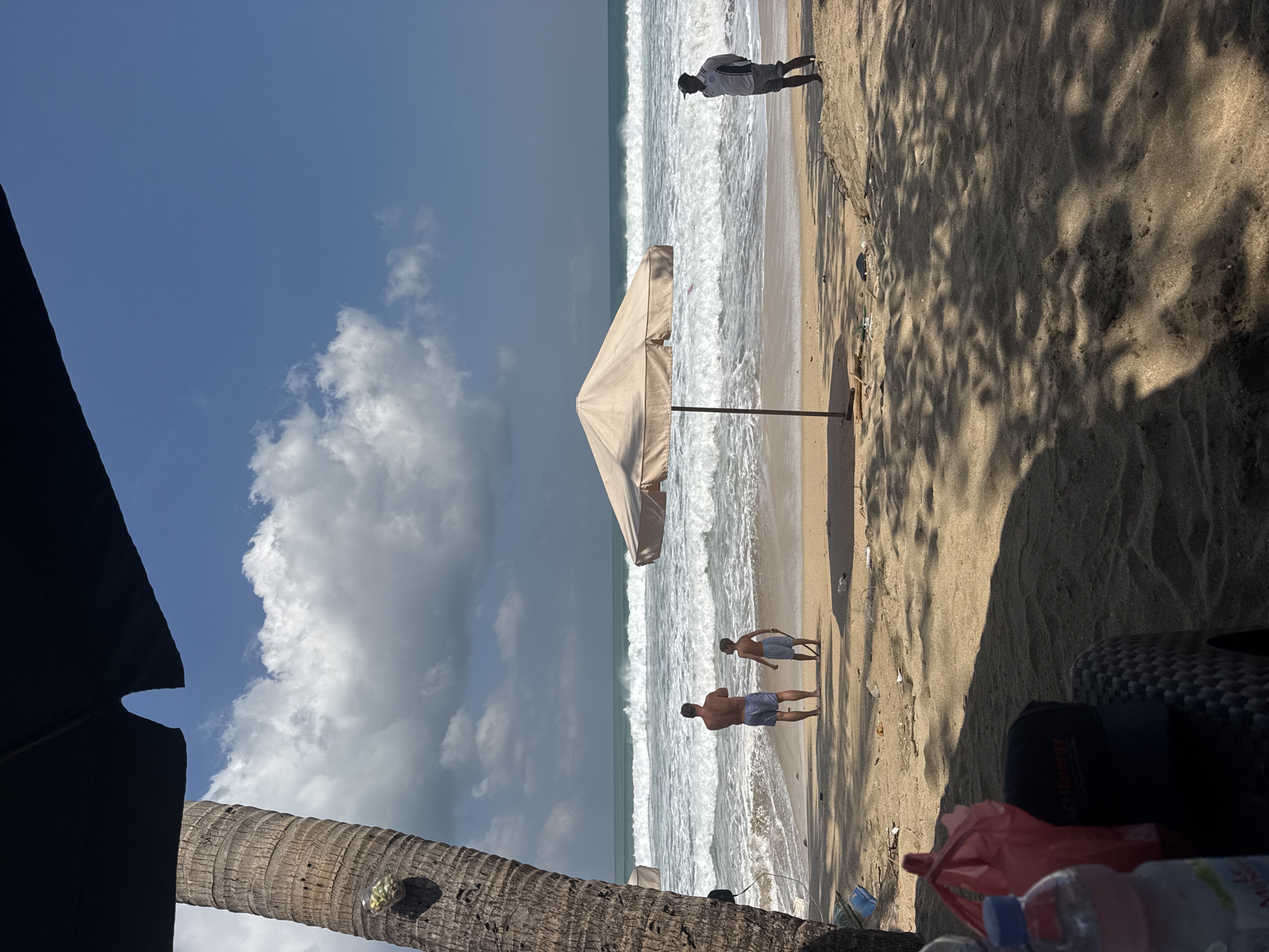 A wide shot captures a serene beach scene in Kuta, Indonesia, under a bright blue sky dotted with fluffy white clouds. The foreground is dominated by the sandy beach, with shadows cast by an unseen umbrella creating a dappled pattern on the sand. To the left, the textured bark of a palm tree stands tall, with a small, clear plastic bag containing what appears to be fruit attached to it. In the middle ground, two people, an adult male and a young boy, stand facing the ocean, appearing to observe the waves. The adult male is wearing blue patterned shorts, and the boy is wearing blue shorts and holding a stick. A large, light-colored beach umbrella stands between them and the water, offering shade. Further to the right, a third individual, a male wearing a dark cap, a light-colored shirt with a dark collar and sleeve trim, and dark shorts, stands facing the ocean, with his hands clasped behind his back. The ocean itself is a vibrant blue-green, with significant waves rolling onto the shore. Debris, including plastic bags, is scattered along the high tide line. In the immediate foreground, a portion of a black woven chair is visible, with a red plastic bag and a partially visible water bottle on it. The scene conveys a peaceful day at the beach.