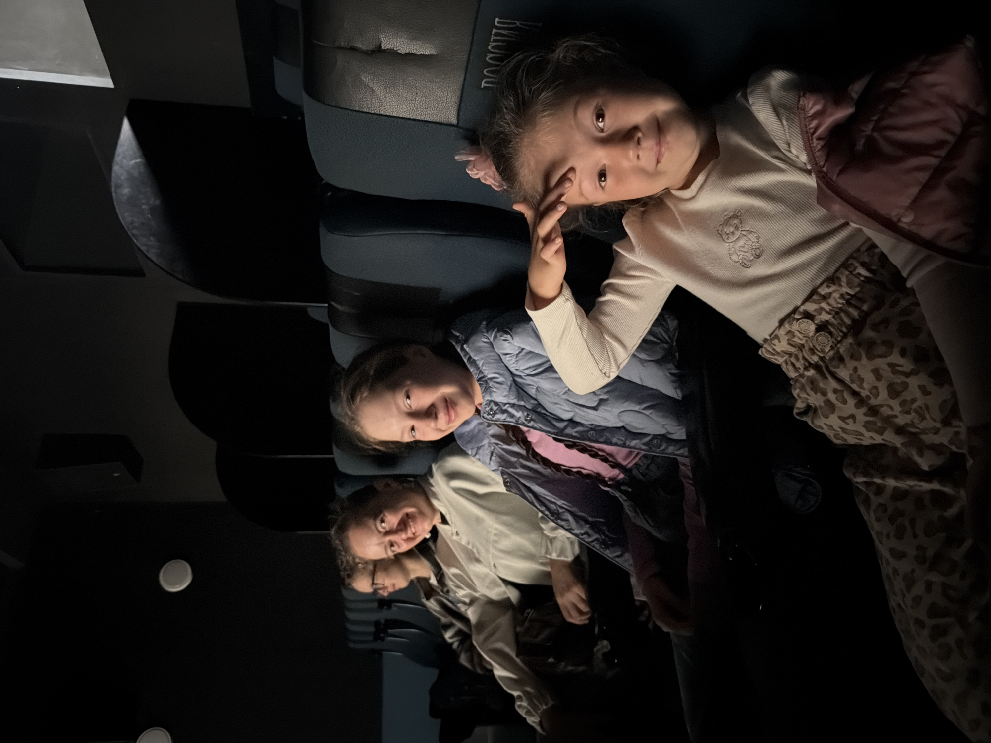 A group of people, likely a family, are seated in a dimly lit cinema. In the foreground, a young girl with a pink bow in her hair smiles at the camera, her hand touching her forehead. She wears a light-colored sweater with a bear emblem and patterned leggings. To her left, another young girl in a blue jacket looks towards the camera with a slight smile. Behind them, a woman with glasses, wearing a white top, is visible. The seats are upholstered in a dark fabric, with some showing wear. The background is dark, indicating the cinema screen is either off or projecting a dark image. The seats have headrests with a textured pattern and embossed Cyrillic text that appears to say "РОССИЯ" (Russia). The overall lighting is low, creating a typical cinema ambiance. The presence of "РОССИЯ" on the seats strongly suggests the location is in Izhevsk, Russia, as this is a common theater chain name in Russia.