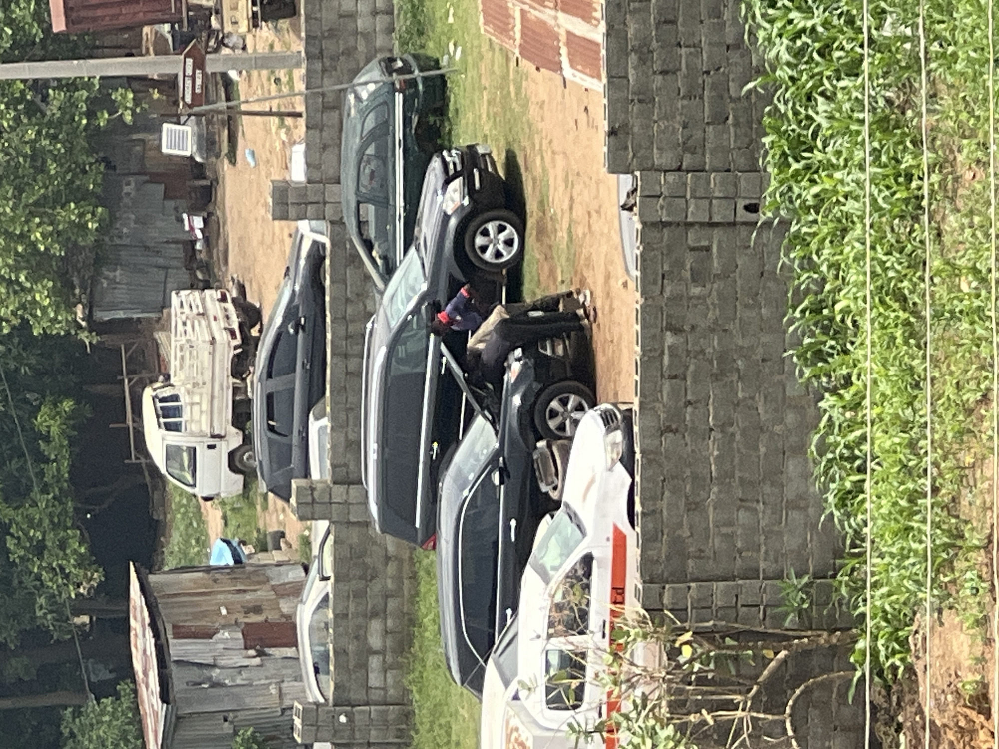 The image depicts a row of parked cars along a wall in Abuja, Nigeria. Several dark-colored SUVs and an ambulance are visible. A man wearing dark clothing and a hat is seated on the side of one of the SUVs. Beyond the cars, there are buildings with corrugated metal roofs and a white van. There is greenery in the foreground and trees in the background. A street sign is attached to a light pole in the distant background. The ground is dirt or gravel.