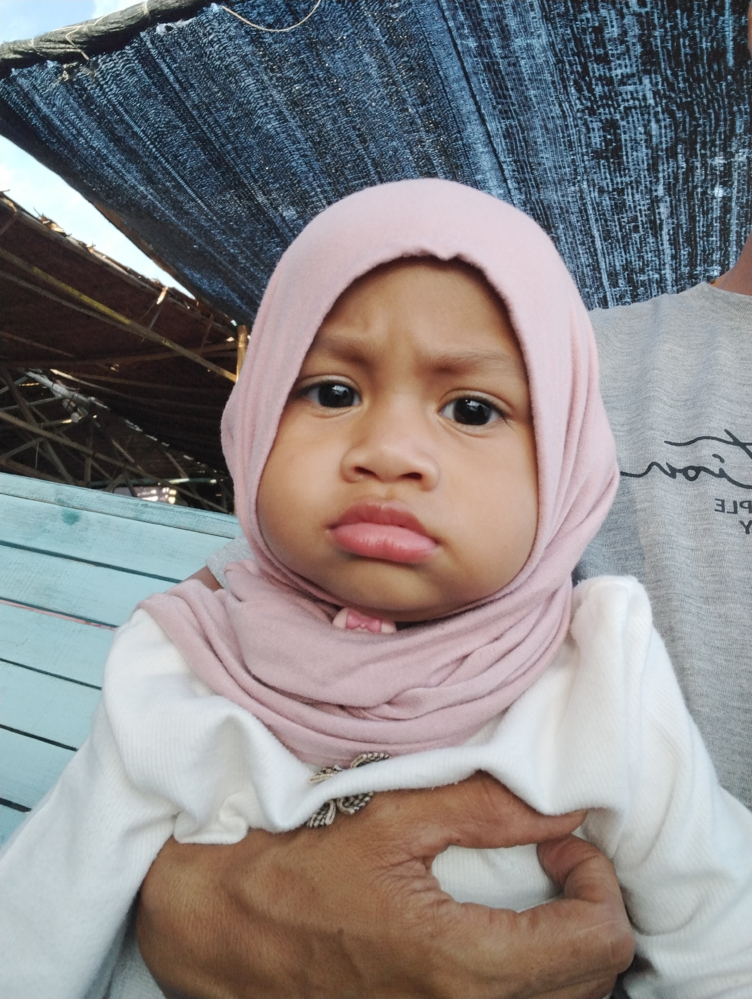 A close-up, eye-level shot shows a young child wearing a light pink hijab and white long-sleeved shirt. The child has a neutral to slightly sad expression. An adult holding the child is visible, with portions of their grey shirt and skin-toned arm in view. In the background, there are sections of light blue wooden slats and a dark, coarsely textured overhead covering. Based on the prompt details provided, the image is likely taken in Lhokseumawe, Indonesia.