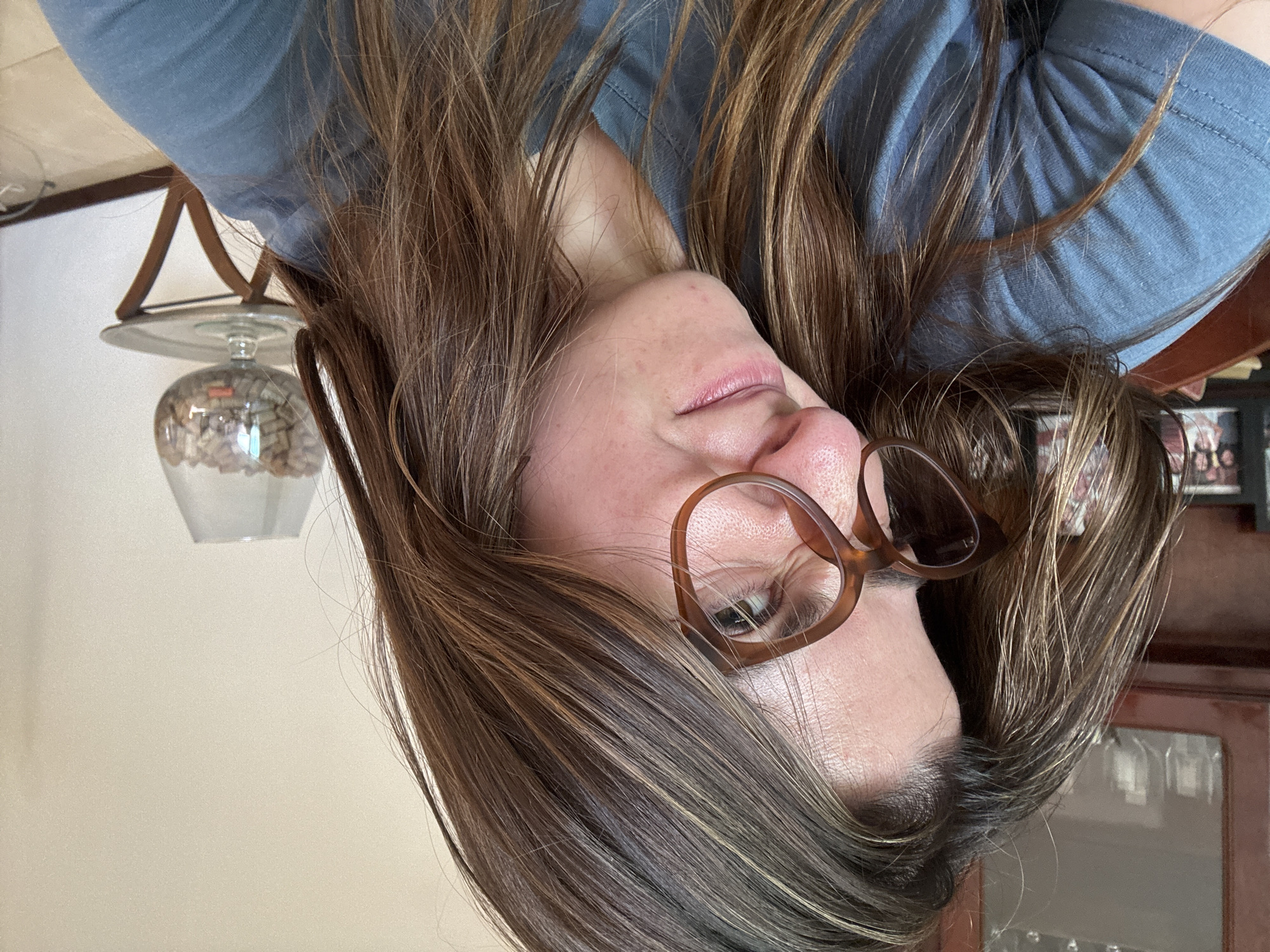 A woman with light brown hair and glasses is centered in the image, tilting her head. She is wearing a blue top. In the background, a lamp sits on a table next to a cabinet.