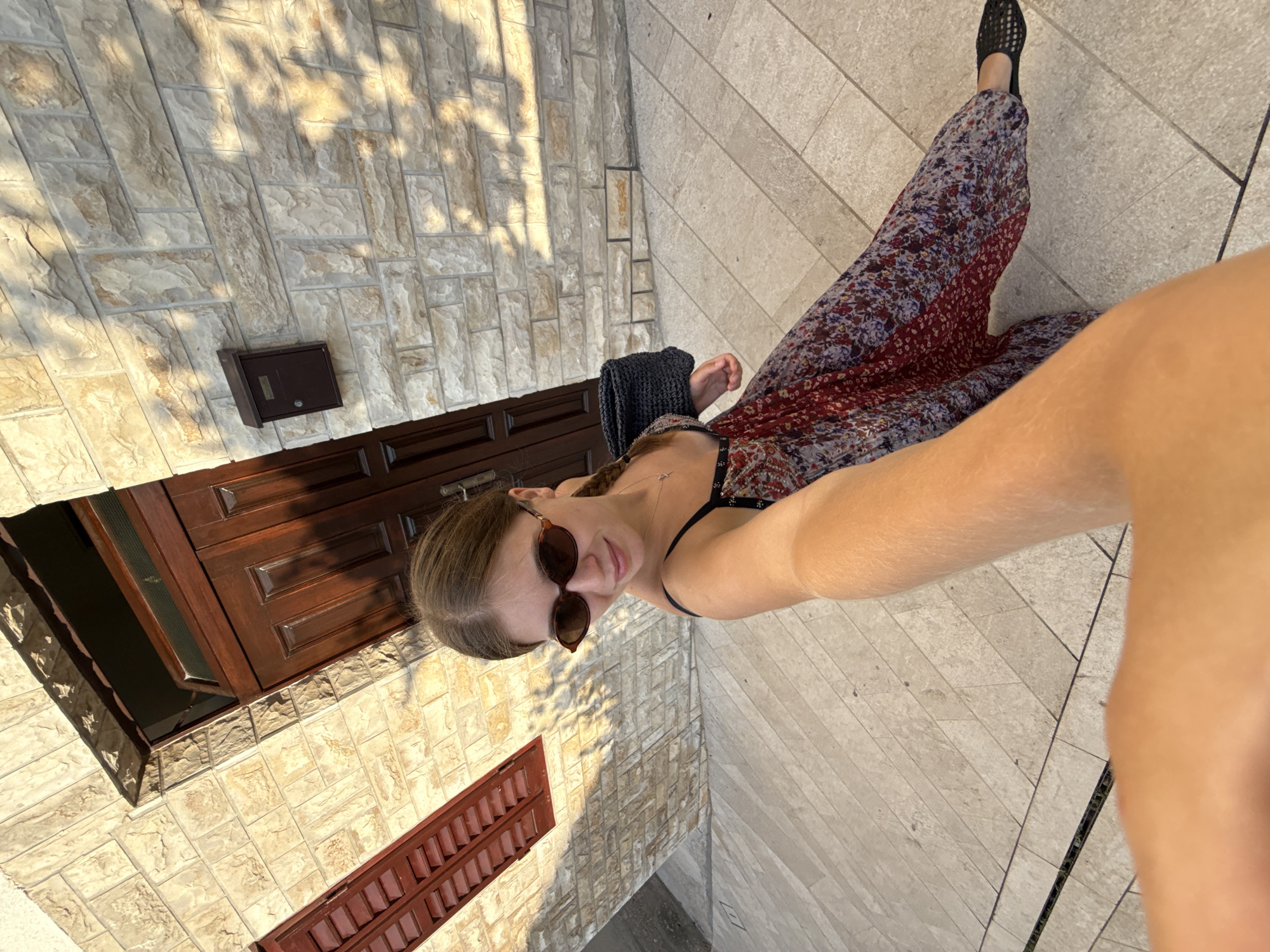 A woman with braided blonde hair and sunglasses takes a selfie outside a stone building in Primošten, Croatia. She is wearing a floral maxi dress and black woven sandals. A black straw bag is slung over her shoulder. The building has a wooden door and window shutters. The ground is paved with large stone tiles. Sunlight casts shadows on the building and ground.