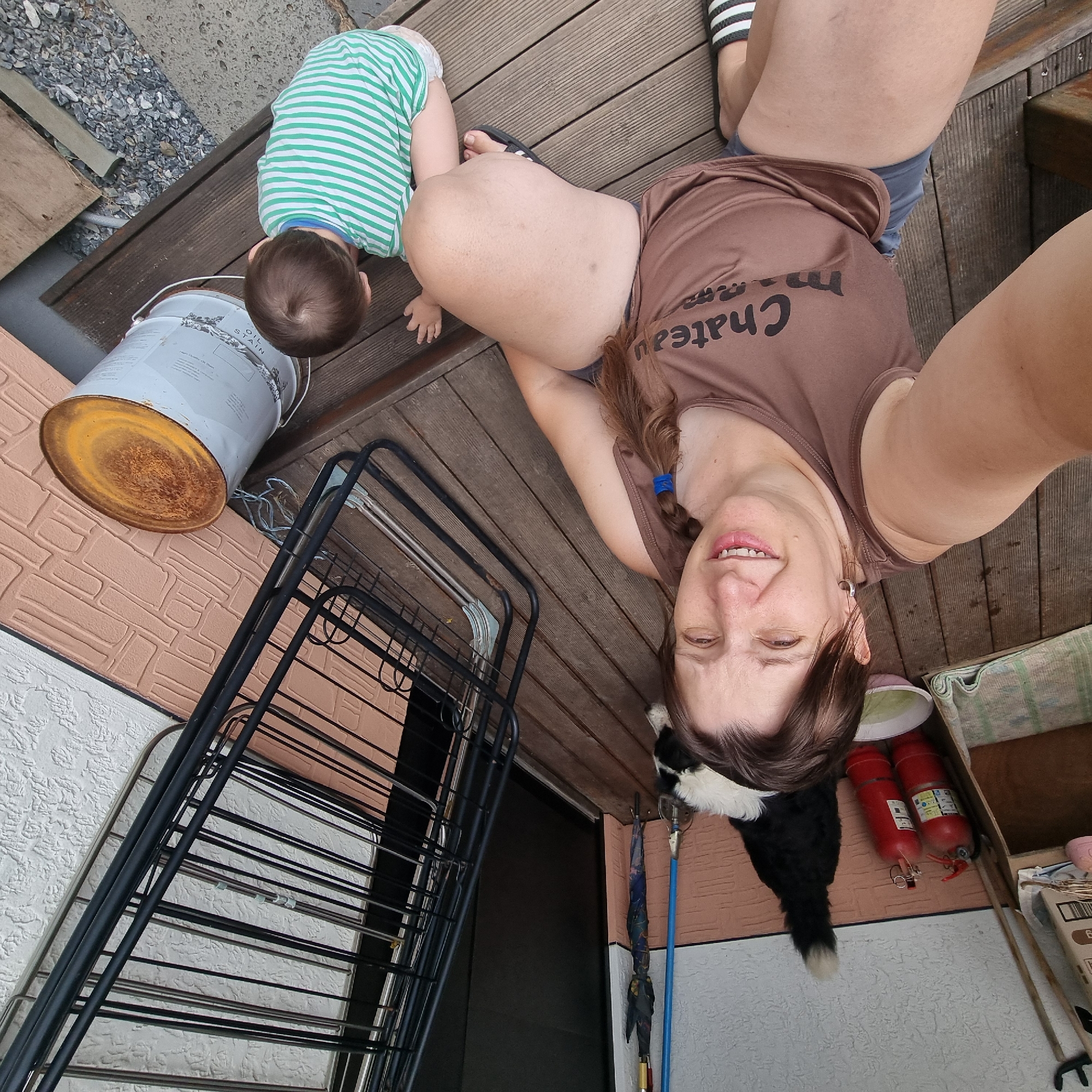 This is a first-person perspective photograph taken on a wooden deck. A woman is in the foreground, smiling at the camera. She is wearing a brown tank top with the words "Chateau maman" printed on it, and blue shorts. Her hair is brown and braided. To her left, a black and white dog is lying down. To her right, a young child in a green and white striped shirt and a diaper is seated on the deck, facing away from the camera. In the background, there are two red fire extinguishers, an umbrella, a drying rack, and a large metal can with a rusty lid. The environment appears to be an outdoor porch or patio.