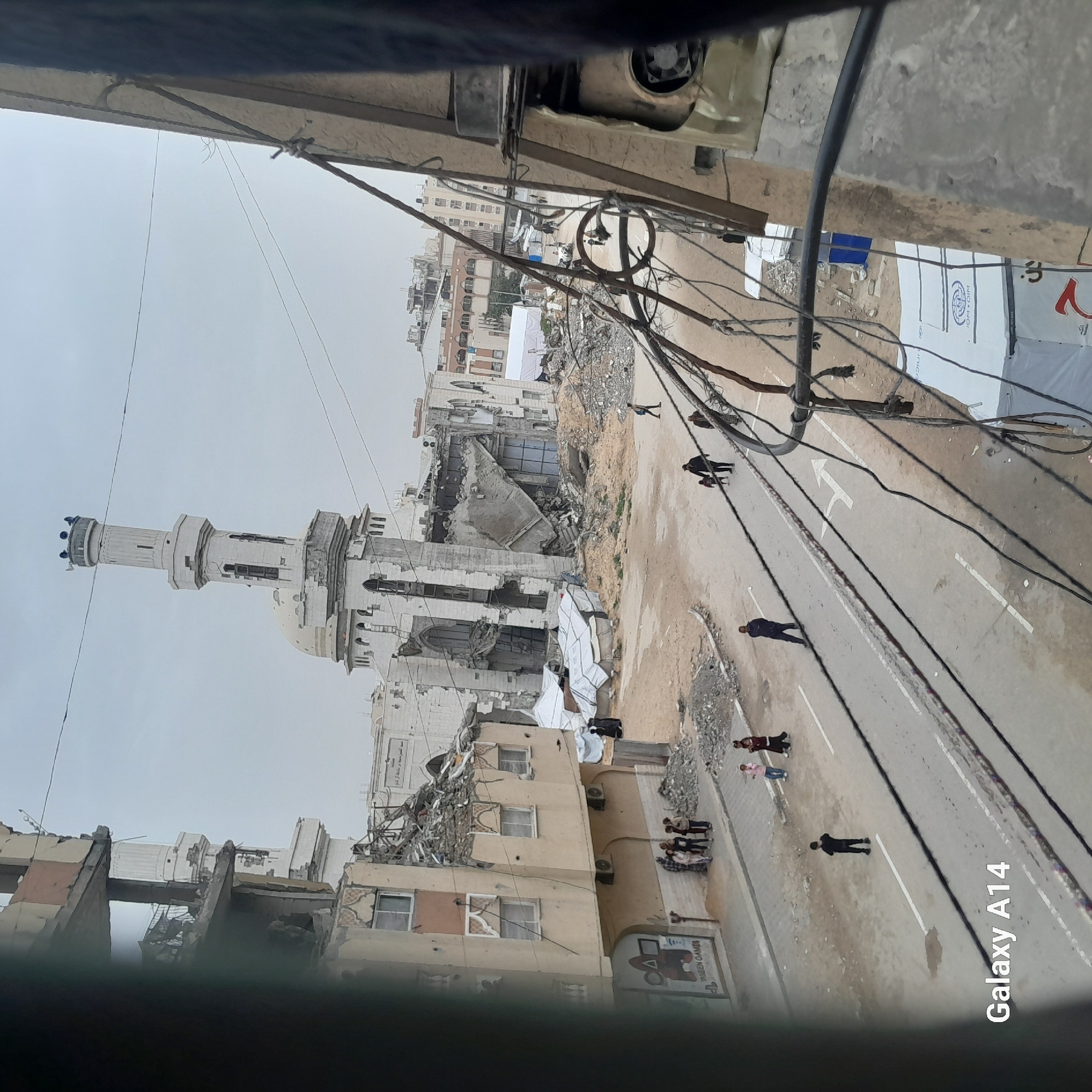 The image depicts a street scene in Khan Yunis, Palestinian Territory, revealing significant destruction. A large mosque with a tall minaret stands amidst partially collapsed buildings, indicating bombing or other conflict-related damage. Rubble is scattered throughout the scene. Several individuals are visible walking along the street, seemingly navigating the aftermath. Overhead, numerous power lines crisscross the view, and a partial view from a higher elevation offers a framed perspective of the situation.