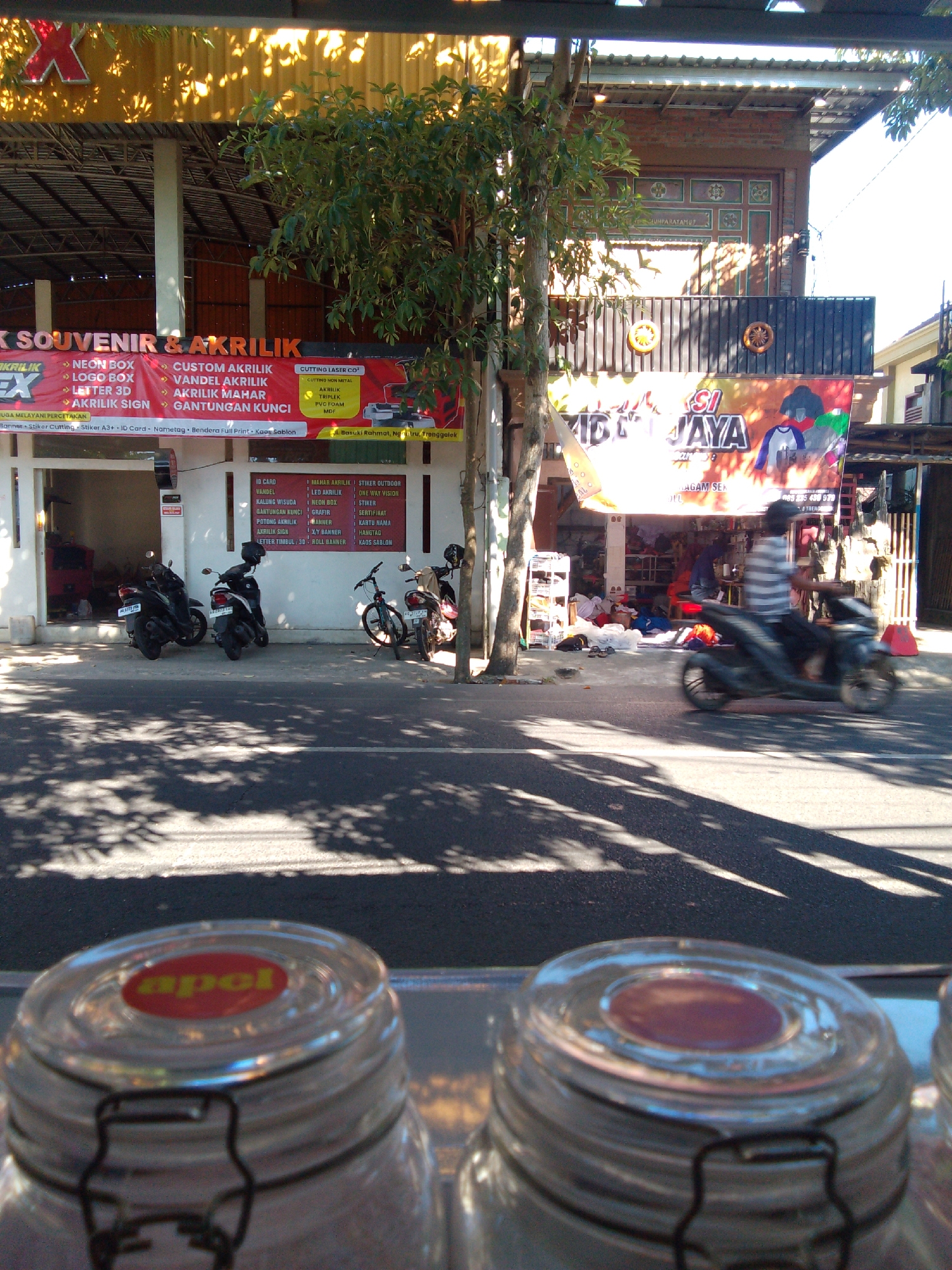 The image depicts a street scene in Trenggalek, Indonesia, likely during daylight. In the foreground, the lids of two glass jars are visible. The scene then moves to a storefront that appears to be a shop specializing in souvenirs and acrylic items. A large banner advertising "SOUVENIR & AKRILIK" is prominently displayed, listing various services such as neon boxes, logo boxes, acrylic signs, and custom acrylic work. Another banner, featuring "SIDJAYA," is also visible, with an image of a shirt. Several motorcycles are parked in front of the shop. A person is seen riding a motorcycle on the road in front of the storefront, captured in motion. The environment includes a street, a sidewalk, and a building with traditional Indonesian architectural elements. The overall impression is that of a commercial area in a city.