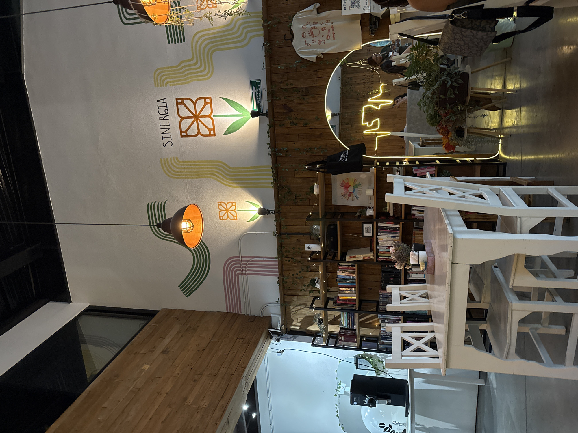 The image depicts the interior of a cafe in Puebla, Mexico. The walls are adorned with painted designs, including floral patterns and the word "SINERGIA." Exposed light bulbs provide illumination. A wooden bookshelf filled with books stands against one wall. Tables and white wooden chairs are arranged for customers. A T-shirt is hanging, a mirror, and a coffee machine are visible. People are sitting at tables, engaged in conversation. The cafe appears to be a casual and inviting space.