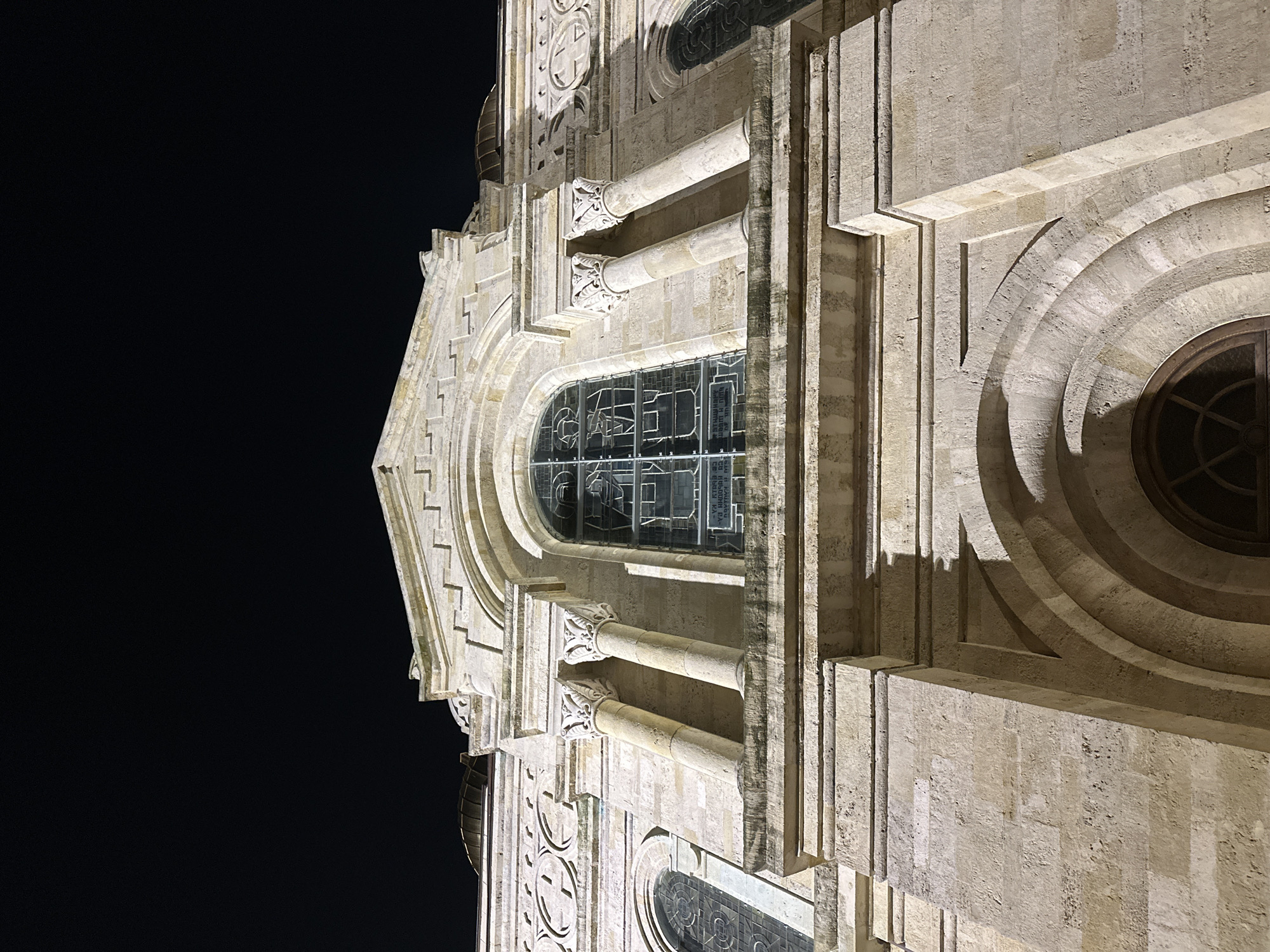 The image depicts a section of a building, specifically the facade of the Dormition of the Mother of God Cathedral in Varna, Bulgaria, taken at night. The architectural style includes light-colored stonework, columns with ornate capitals, arched windows with leaded glass, and circular windows with concentric rings. The building is illuminated by artificial light, which emphasizes its details against the dark night sky. There are no people visible. The facade's intricate stonework and architectural elements are prominently featured. The overall composition conveys a sense of grandeur and historical significance.