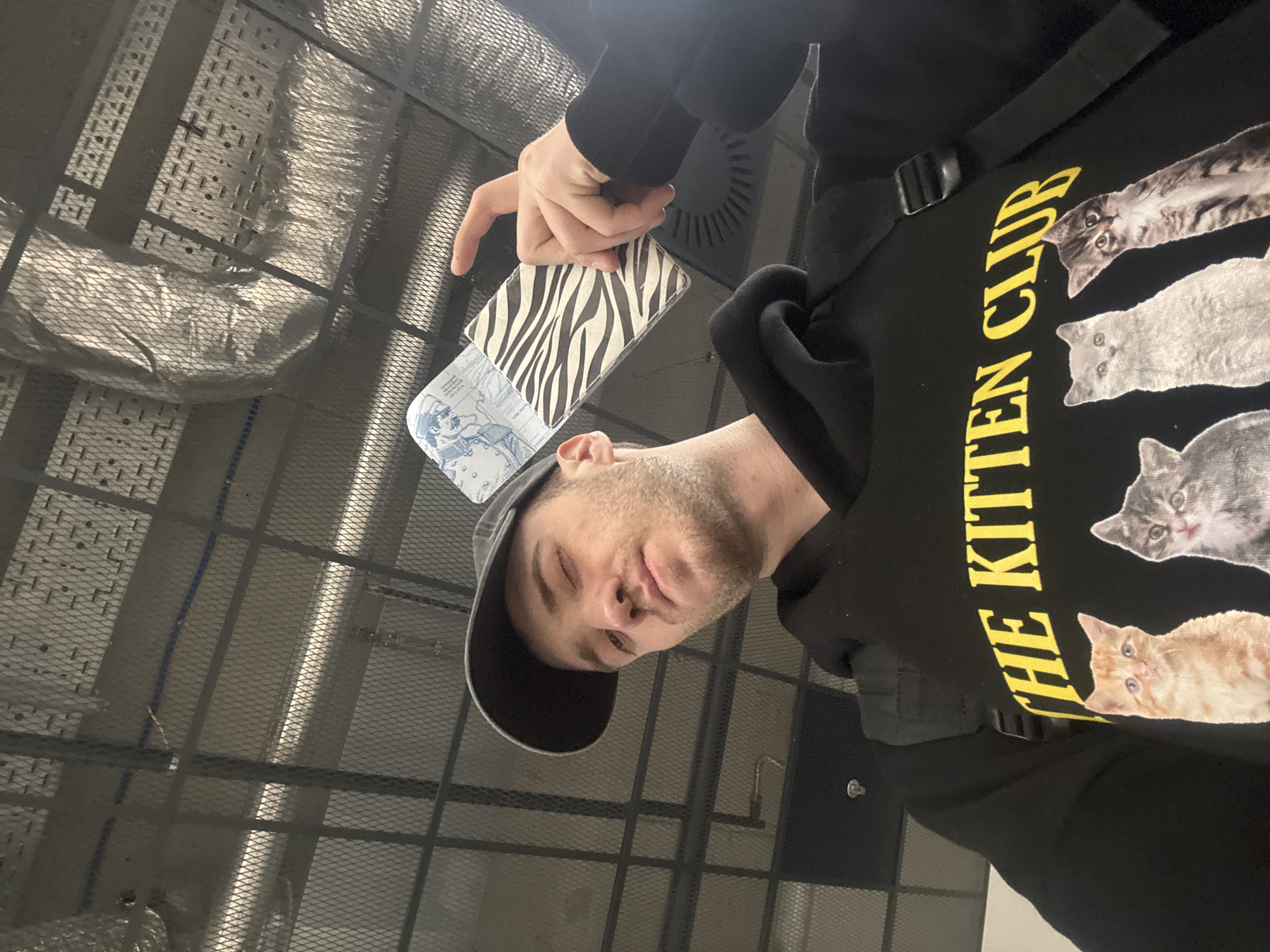 A man wearing a black baseball cap and a black sweatshirt with a print of cats and the words "THE KITTEN CLUB" is holding a passport with a zebra-print cover. The passport appears to be open, displaying a picture of a man in uniform. The setting appears to be indoors, with a metal grid or grating structure visible in the background. Given the passport’s image, the city of Khabarovsk in Russia can be the location.