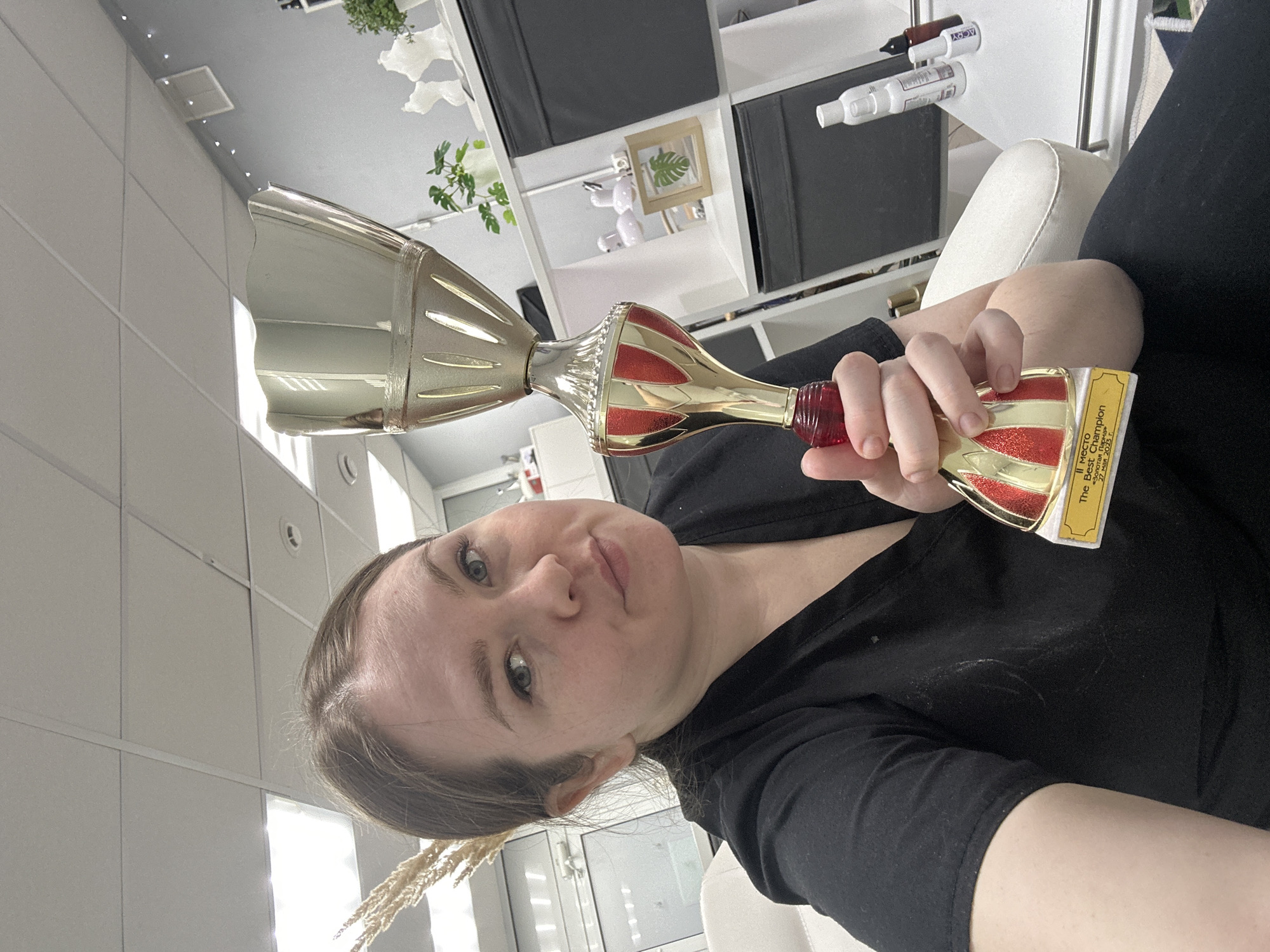 A woman is holding a gold trophy with red accents and a label written in Russian that reads "II место The Best Champion г. Чайковский 27 мая 2023 г.". The woman is wearing a black shirt and is sitting in front of a white wall with shelves and plants. The location is Chaikovsky, Russia.